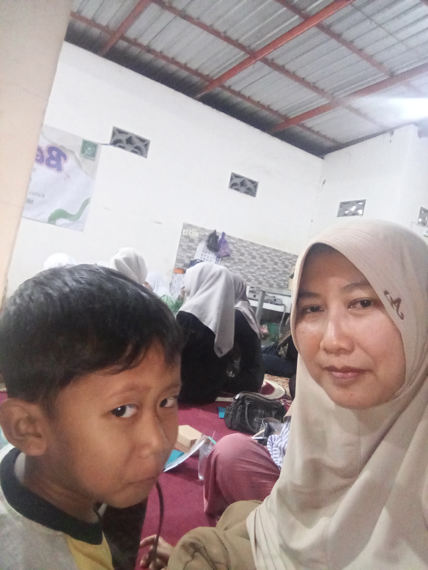 A close-up selfie taken indoors shows a young boy in the foreground, his face turned towards the camera with a slightly worried expression. Behind him, a woman wearing a light-colored hijab is also looking at the camera. In the background, other people are visible, some wearing hijabs, sitting on the floor. The room appears to be a simple indoor space with a corrugated metal ceiling and white walls. There are ventilation blocks visible on the walls. A banner with the letter "B" prominently displayed and a green emblem is affixed to one of the walls. The flooring is a reddish-pink carpet. This scene is located in Mlilir, Indonesia.