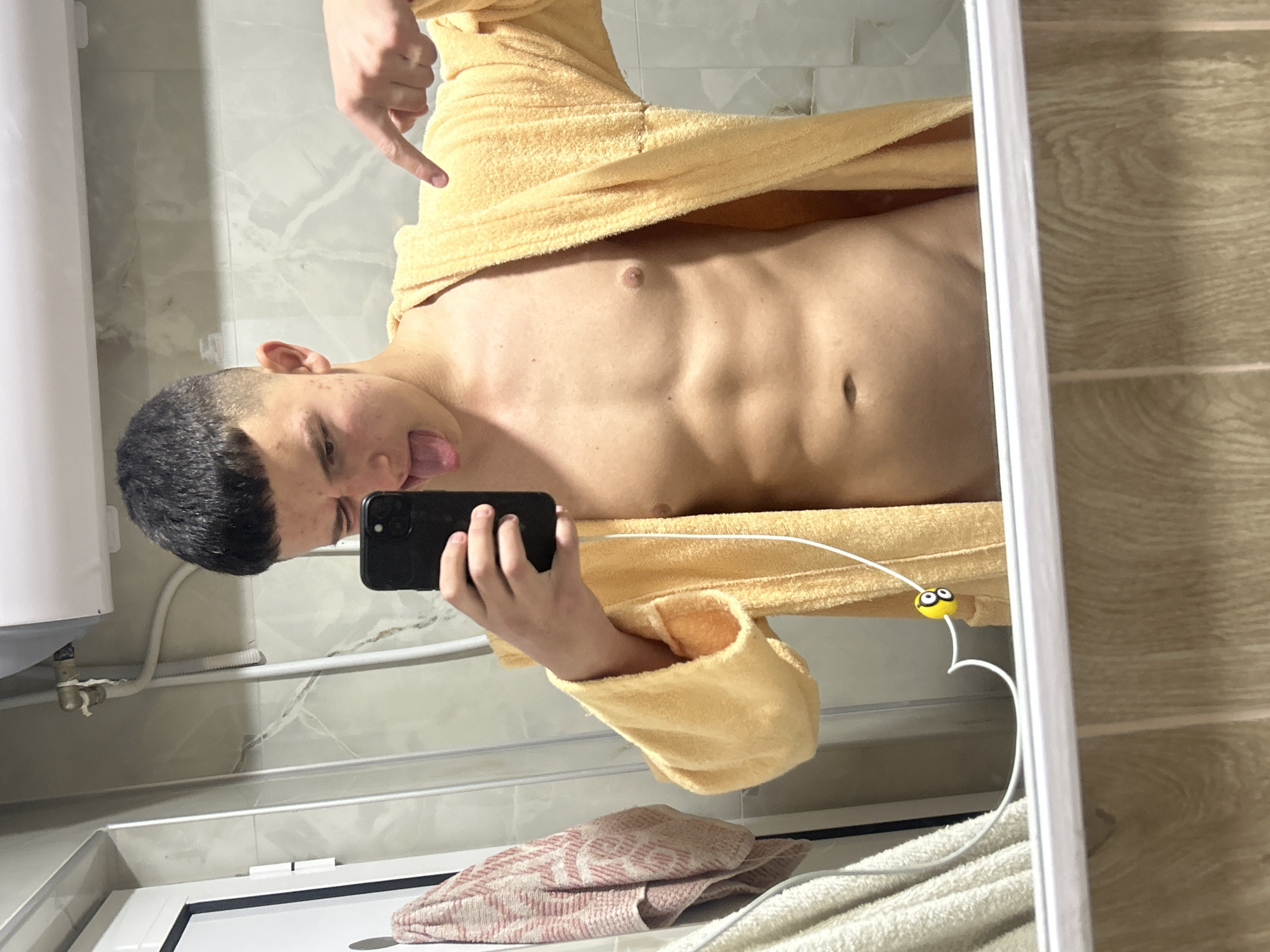 A young male individual is depicted taking a mirror selfie in what appears to be a bathroom. He is shown from the mid-chest upwards, with his head slightly tilted and his tongue playfully sticking out. He has short dark hair and visible blemishes or acne on his face. He is wearing a yellow or tan terry cloth bathrobe, which is open, revealing his toned abdomen with visible muscle definition and a navel piercing. His left hand holds a black smartphone, capturing the reflection, while his right hand is raised with the index finger pointing downwards towards his torso.
The background, reflected in the mirror, features light gray tiled walls with a subtle marble-like pattern. To the far left, a white water heater or similar appliance is visible, with some plumbing pipes beneath it. The right side of the background shows wooden-patterned wall or floor tiles. In the foreground of the reflection, below the individual, there's a sink area or countertop where two folded towels, one pink patterned and one plain light-colored, are placed. A white cable, possibly for charging, with a small yellow "Minion" character charm attached, is draped over the bathrobe. The overall impression is casual and playful, taken indoors.