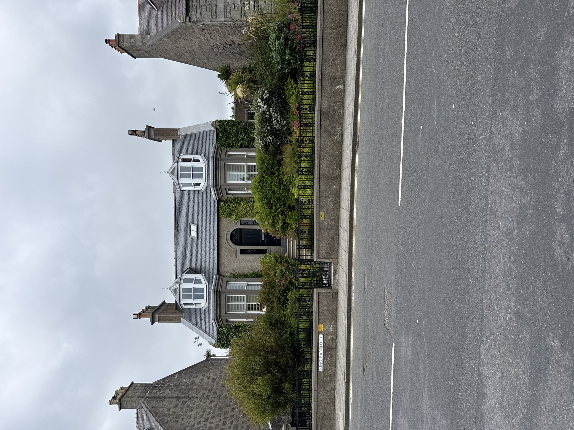 The image depicts a detached house situated on King Harald Street in Lerwick, United Kingdom. The house, characterized by its grey stone facade and slate roof, features prominent bay windows on the front, adorned with white frames. A black front door is centrally located, accessible via a small set of steps. The property is enclosed by a black wrought-iron fence and a low stone wall, behind which a lush garden with various plants and flowers is visible. The street in front of the house is asphalted, with white painted lines indicating a road. The sky is overcast, suggesting cloudy weather. The scene is devoid of any individuals or discernible activity.