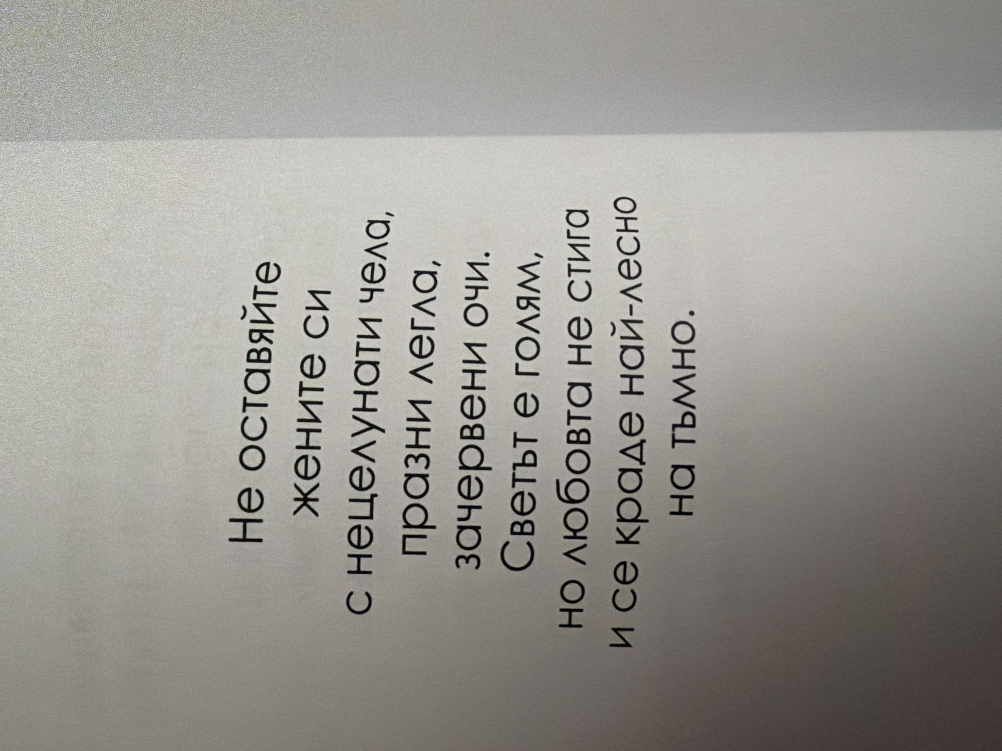 The image shows a page from a book with Bulgarian text printed in black. The text is arranged in several lines, centered on the page. The background is a light, textured surface, possibly paper. There is a subtle gradient in the background, with the right side appearing slightly darker. The lighting appears to be coming from the top left.
The text reads:
Не оставяйте
жените си
с нецелунати чела,
празни легла,
зачервени очи.
Светът е голям,
но любовта не стига
и се краде най-лесно
на тъмно.
The translation of the text is:
"Do not leave
your women
with unkissed foreheads,
empty beds,
reddened eyes.
The world is big,
but love is not enough
and is stolen most easily
in the dark."
There are no people, subjects, objects, or actions visible in the image, other than the printed text itself. The environment is limited to the page of the book and its immediate surroundings, which appear to be a neutral, indoor setting. There is no visible connection to the city of Pazardzhik, Bulgaria, or any specific location within it.