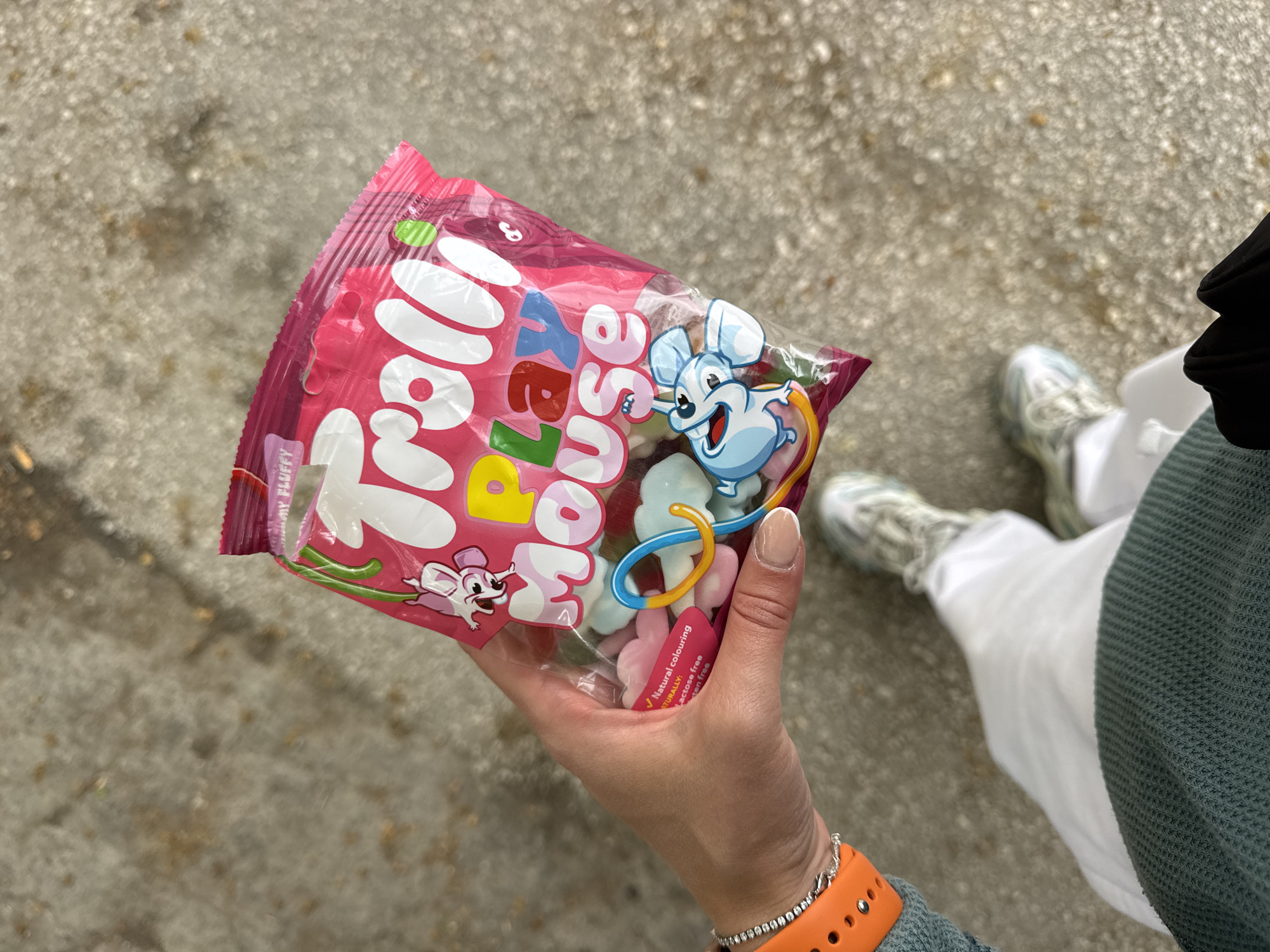 An overhead, close-up shot captures a person holding a vibrant pink bag of "Mousie Mousse" candies outdoors. The hand, with neatly manicured, light-colored nails, holds the transparent bag, showcasing the colorful, soft-looking candies within. The candy pieces are small, cloud-like or mouse-shaped, in shades of light pink, white, light blue, and yellow.
The packaging prominently features the brand name "Mousie Mousse" in large white, bubbly letters, accompanied by cheerful cartoon mice—a larger blue one with a yellow cheese-like loop and a smaller pink one. Additional text on the bag includes "FLUFFY" and partially visible claims like "Natural colouring," "Lactose free," and "Gluten free."
In the background, parts of the person's attire are visible: a white pant leg, a light-colored sneaker (possibly grey and white), and a textured green or teal garment on the upper right, likely a sleeve. The wrist holding the candy bag sports a bright orange watch band and a delicate silver-colored bracelet.
The scene is set on a textured, grey concrete or paved surface, indicating an outdoor environment, possibly a sidewalk. The lighting is soft and even, suggesting daytime without harsh sunlight. The activity appears to be a casual moment of holding or enjoying the candy. The location context is Blagoevgrad, Bulgaria.