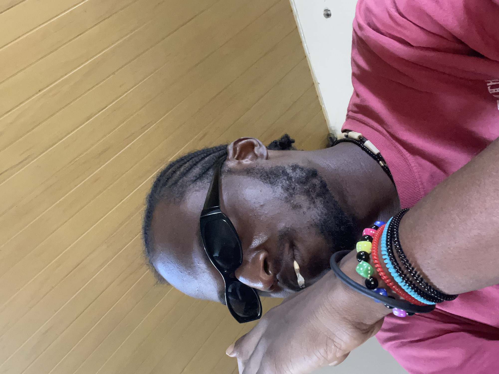 A Black male is depicted in an indoor setting, observed from a low-angle perspective looking upwards and to his left. He has a dark complexion, a short beard, and braided hair. He wears black, wrap-around sunglasses that obscure his eyes. A hand-rolled cigarette, consistent with a joint, is positioned between his lips on the right side of his mouth, appearing lit at the tip. He is dressed in a plain, short-sleeved, bright pink t-shirt and wears a multi-strand necklace composed of dark and light-colored beads. His left forearm and wrist are prominent in the foreground, adorned with a thick black band, a beaded bracelet featuring colorful dice-shaped beads (green, yellow, pink, blue, purple), and several coiled bracelets in red, light blue, and black. His left hand is raised towards his face. The background consists of a light brown wooden paneled ceiling, arranged diagonally, and a section of a white wall in the lower right. This scene takes place in Jos, Nigeria.
