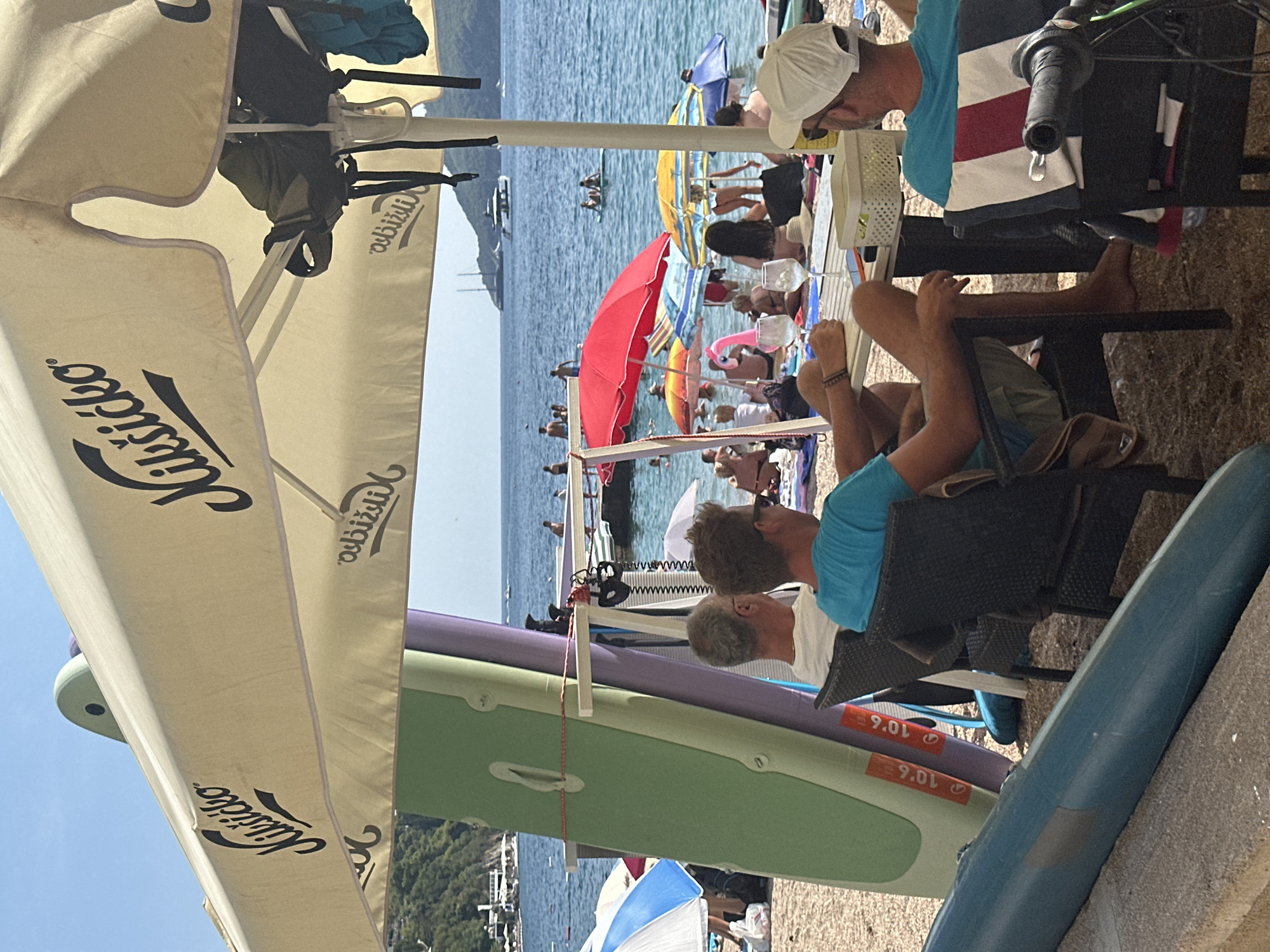 The image depicts a crowded beach scene in Budva, Montenegro, on a sunny day. Several people are seated at tables under a large umbrella, some of whom are being served drinks. In the background, the turquoise waters of the Adriatic Sea are visible, dotted with boats and swimmers. Beach umbrellas and lounge chairs are scattered across the sand, and inflatable toys add a playful element to the scene. The atmosphere appears relaxed and festive, with people enjoying the summer weather and the amenities of a beachside establishment. The presence of paddleboards suggests water sports are readily available.