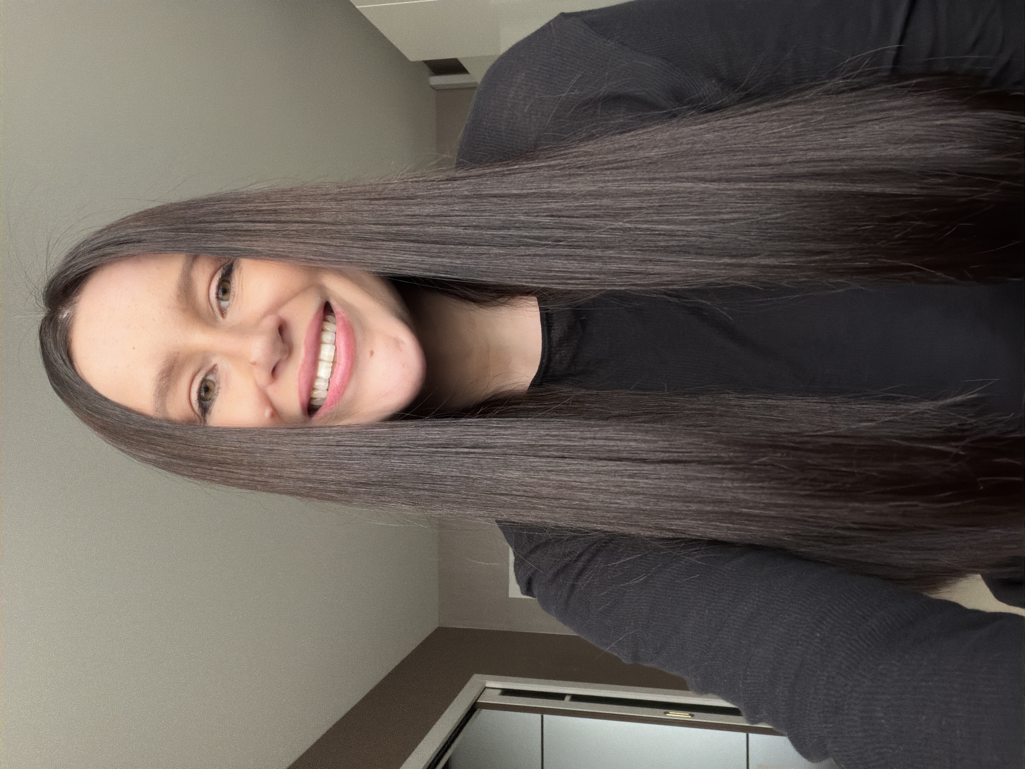 An indoor portrait shows a young woman with very long, straight, dark brown hair, smiling broadly at the camera. The image is oriented on its side, making her hair drape diagonally across the frame. She has light brown eyes and a cheerful expression, revealing white teeth. She is wearing a plain black long-sleeved top. The background consists of two different colored walls: a lighter, neutral gray or beige section in the upper part and a darker brown or taupe section below, possibly a lower wall panel or dado rail. A small portion of what appears to be a doorframe or mirror is visible at the bottom of the frame. The lighting is soft and even, suggesting an indoor setting with ambient light. No visible text is present in the image. Her prominent, healthy-looking hair is a key feature of the photograph.