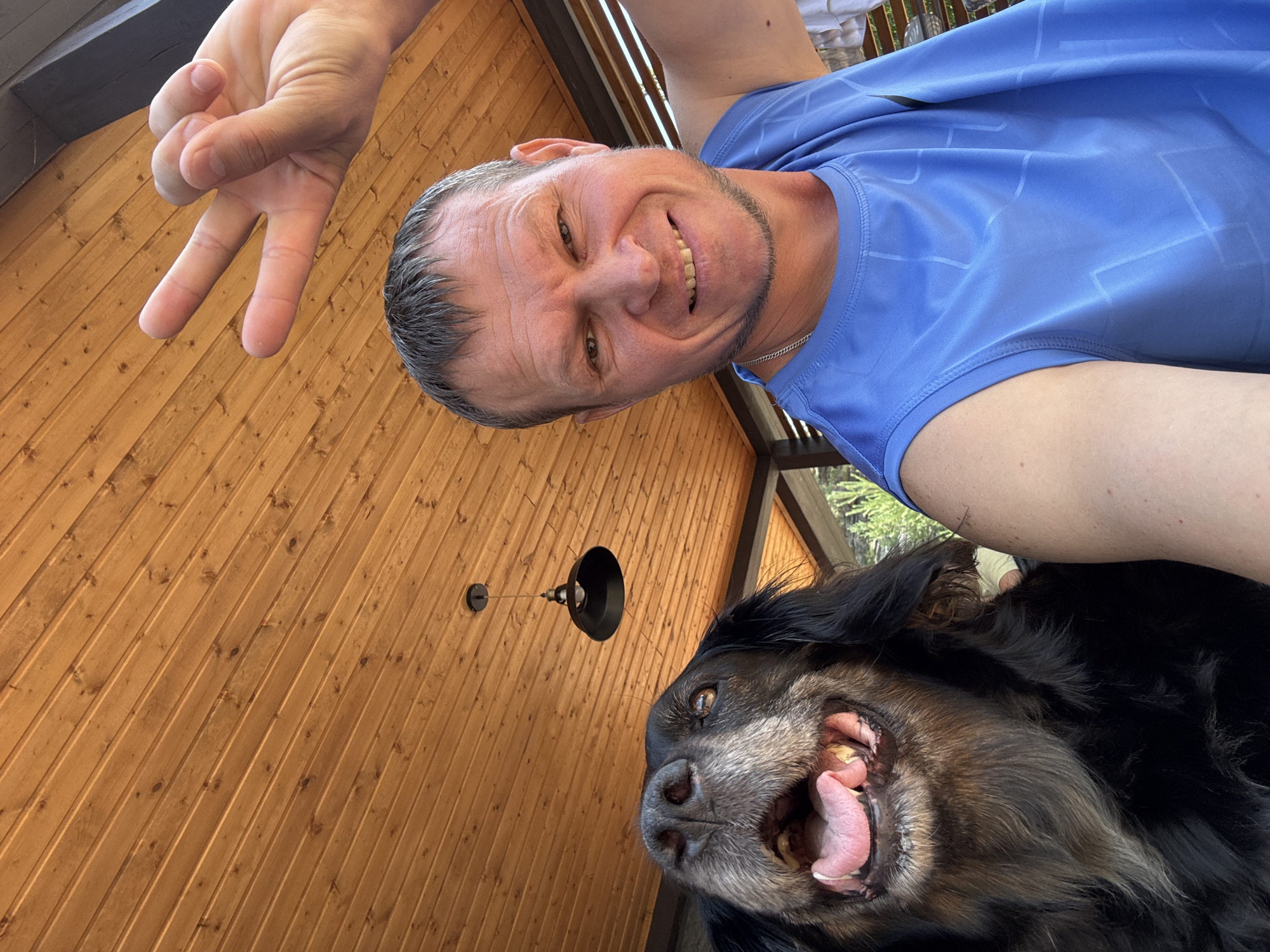 A man and a dog are captured in a selfie taken outdoors under a wooden slatted ceiling, possibly on a porch or under a gazebo. The man, in his late 40s to early 50s, has short, graying hair and is wearing a sleeveless blue shirt. He is smiling and holding up two fingers in a peace sign with his right hand. To his left, a large black dog with its tongue hanging out is looking up at the camera. The dog has graying fur around its muzzle and appears to be a mixed breed, possibly with some retriever or shepherd in it. A black industrial-style pendant light hangs from the ceiling in the background, and the overall lighting suggests it is daytime. The environment appears to be rustic, with the natural wood prominently featured. The man's attire and the outdoor setting suggest a casual, relaxed atmosphere. The image does not provide any specific visual cues that would pinpoint the location to New Bukhtarma, Kazakhstan; it is a generic outdoor setting.