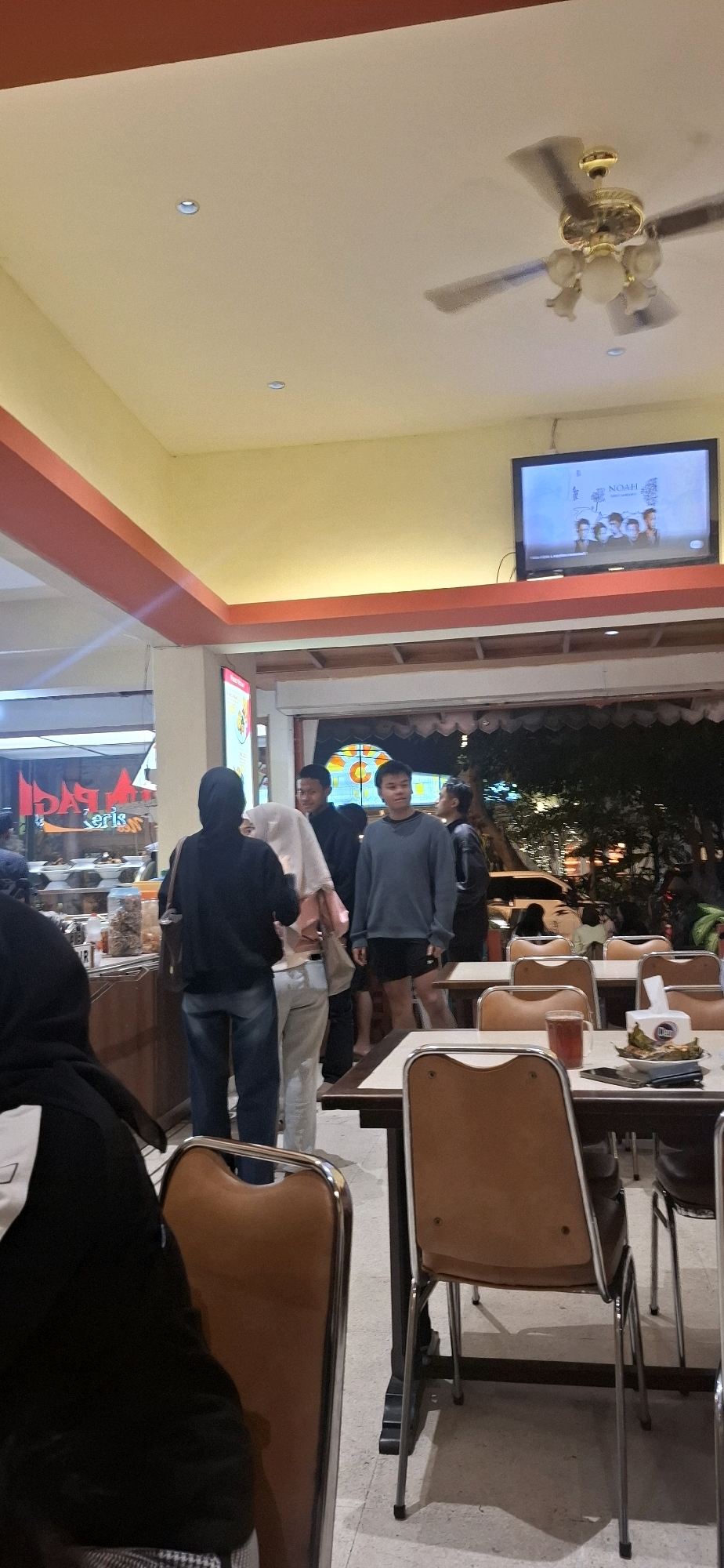 This is a medium shot, eye-level, indoor photograph of a group of people in what appears to be a restaurant or food stall in Surakarta, Indonesia. The lighting is warm and ambient, with some artificial light sources visible, including a television mounted on the wall displaying the word "NOAH" and a group of people. A ceiling fan is also visible overhead, suggesting a warm climate.
In the foreground, on the left side of the image, a person wearing a black hijab and jacket is partially visible, seated at a table. In the center of the image, a group of four people are standing. The person closest to the viewer is wearing a black hijab and a black long-sleeved shirt, with blue jeans. To their right is a person wearing a light pink hijab and a matching shirt, with light-colored pants. Next to them are two men. The man on the left is wearing a dark shirt, and the man on the right is wearing a grey long-sleeved shirt and black shorts.
The environment appears to be a casual dining establishment. Tables and chairs with brown upholstered seats and chrome legs are arranged in the space. On the tables, there are food items, drinks, and a tissue dispenser. In the background, more tables and chairs are visible, along with shelving units that may hold food or other merchandise. Through an open doorway in the background, the exterior of the establishment is visible, showing trees and an outdoor seating area illuminated by artificial lights. The overall impression is that of a busy, casual eating place.