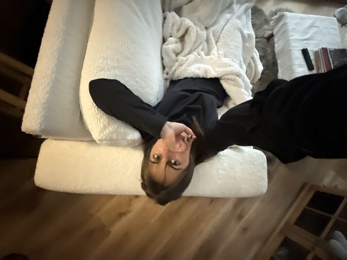 A person lies upside down on a white, fluffy couch with a white blanket. They wear a black top. Dark hair frames their face. A wooden floor is visible, along with a small side table holding a remote and several books or magazines. A wooden cabinet is also present in the background.