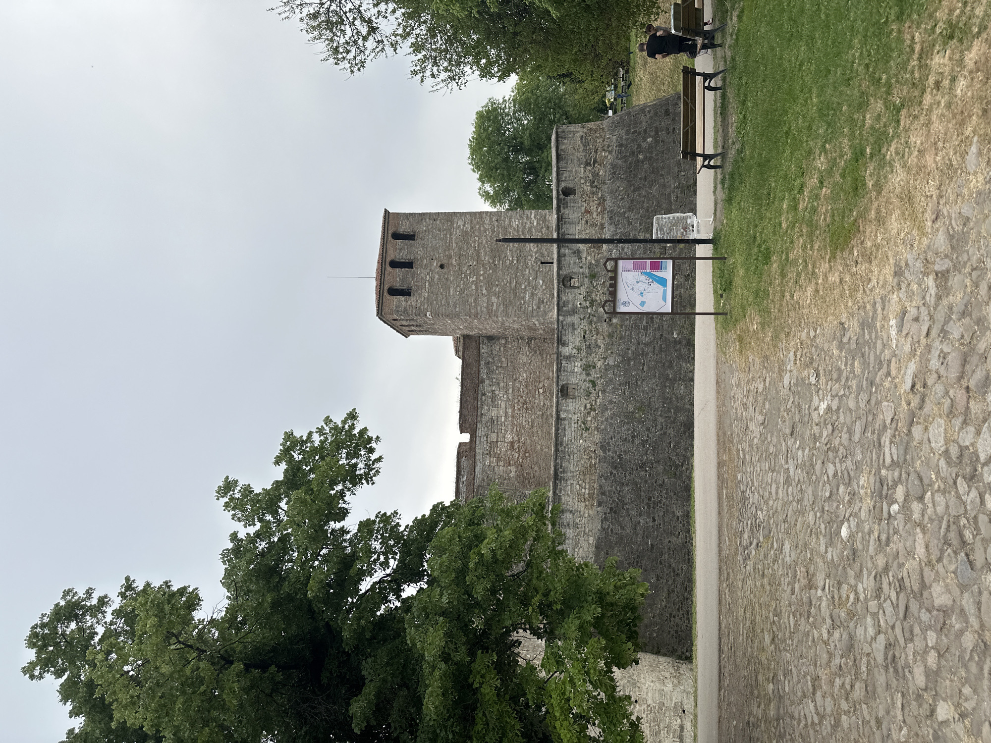 The image depicts a stone tower connected to a wall, possibly part of a fortress or castle, in Vidin, Bulgaria. A signpost stands in front of the wall and tower, with a map or information displayed on it. Adjacent to the wall is a paved pathway. A patch of grass is visible, and trees flank the left side of the scene. In the upper right corner, two individuals sit on a bench. The sky is overcast. The cobblestone path is also notable.