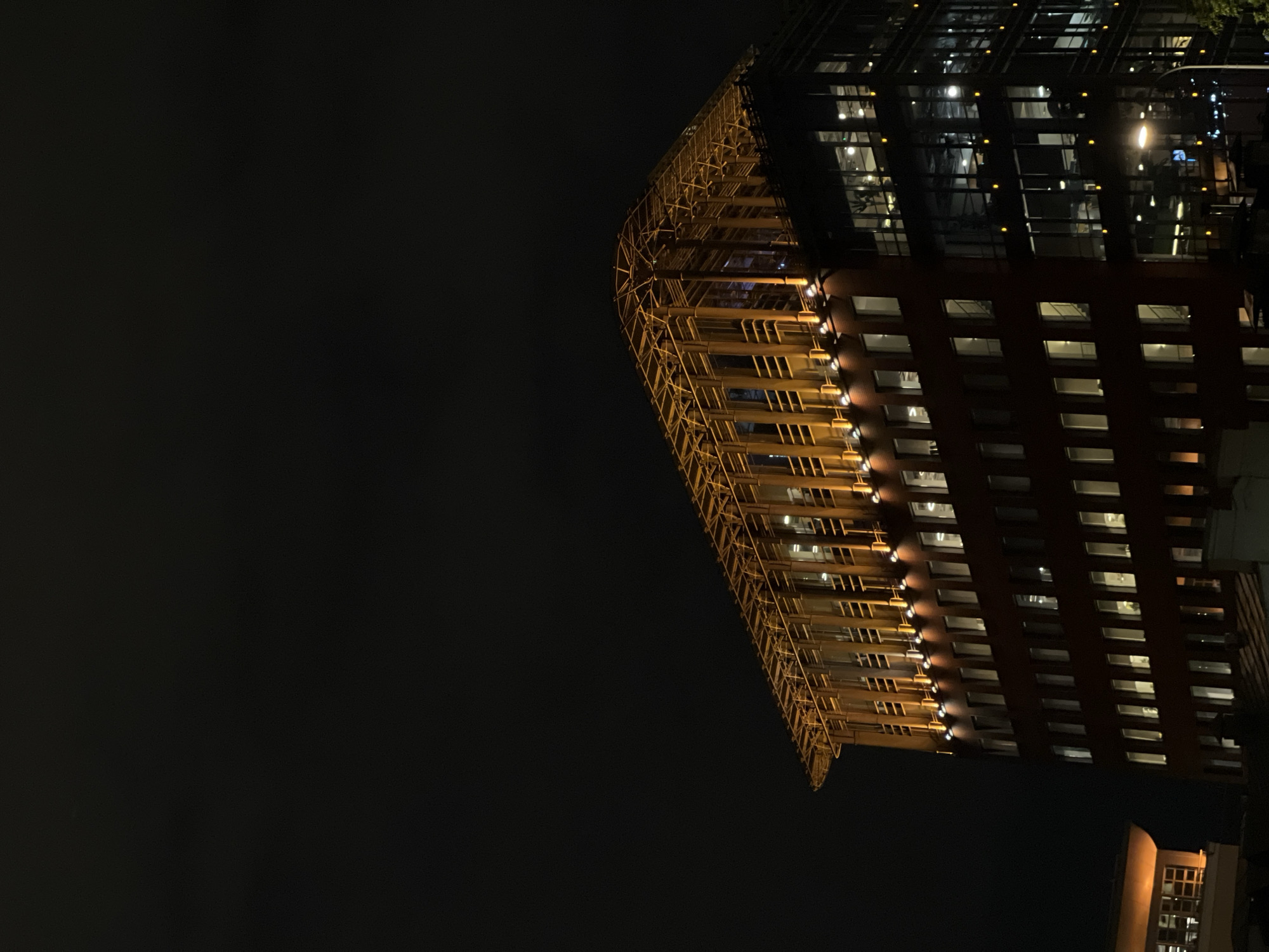The image shows a tall building at night in Moscow, Russia. The building has a distinctive curved facade on one side with exposed metal scaffolding. The rest of the building has a grid-like pattern of windows. Lights are visible inside some of the windows. The night sky is dark and mostly featureless. Another building is visible in the lower right corner of the image, also lit up with windows.