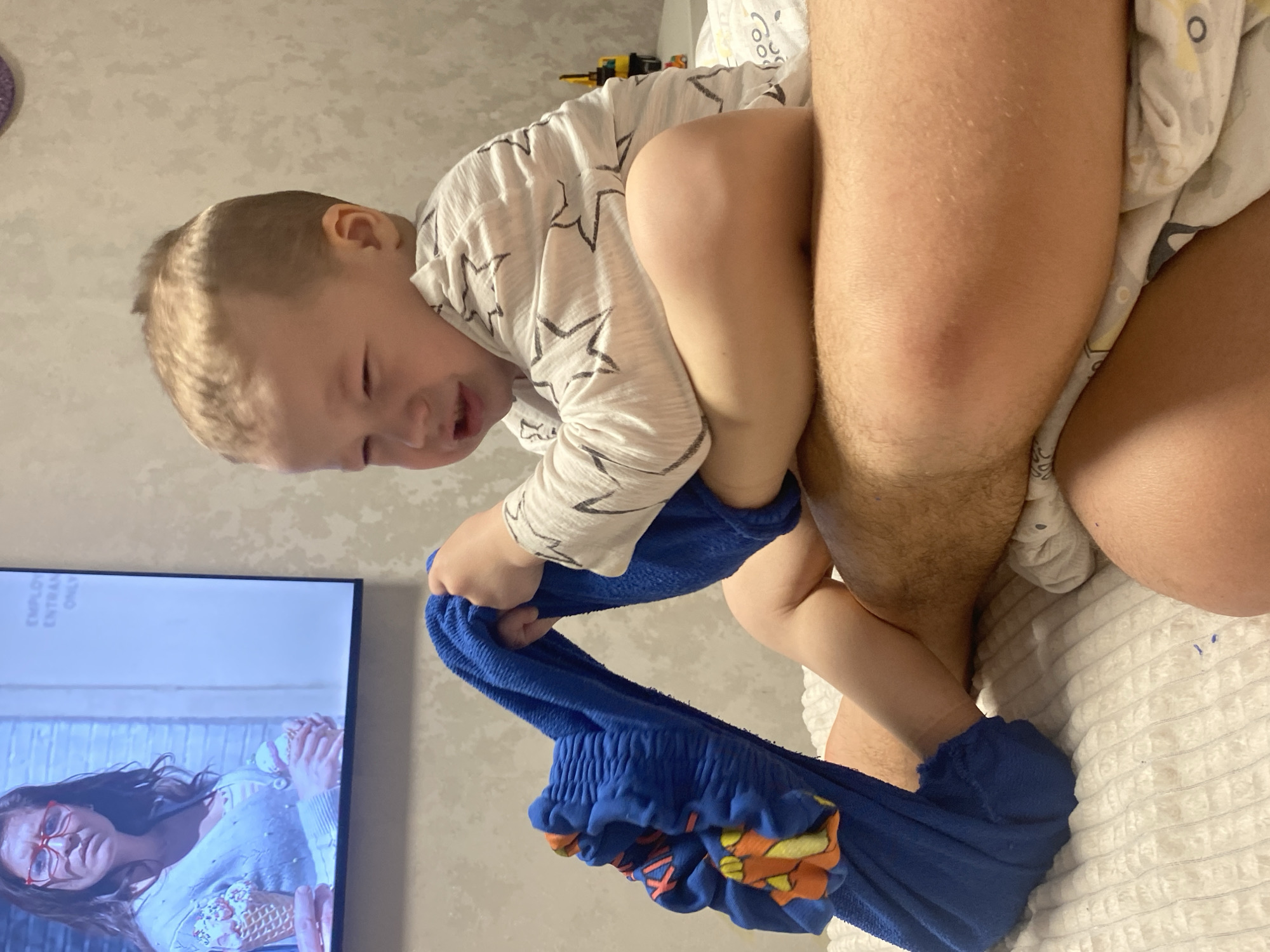 The image depicts an indoor scene, likely a residential setting, without specific geographical markers to pinpoint a location within the city of Sochi, Russia. A light-skinned toddler, approximately 1-3 years old, with short light-brown hair and wearing a white long-sleeved shirt with black star patterns, is seated with crossed legs on the bare, hairy legs of an adult. The child's face is visibly distressed, with eyes squeezed shut and mouth open, as both hands pull on a blue, textured sock adorned with an orange and yellow cartoon fish character. The adult's right thigh, visible beneath the child, exhibits a reddish, slightly raised area. In the background, a flat-screen television is mounted on a textured, light-colored wall, displaying a female individual with dark hair and red glasses, holding two ice cream cones. Partially visible English text on the screen reads "EMPLO" and "ENTRAN ONLY". A yellow and black toy is partially visible on a surface behind the child to the right. The subjects are resting on a white, quilted-patterned duvet or blanket.