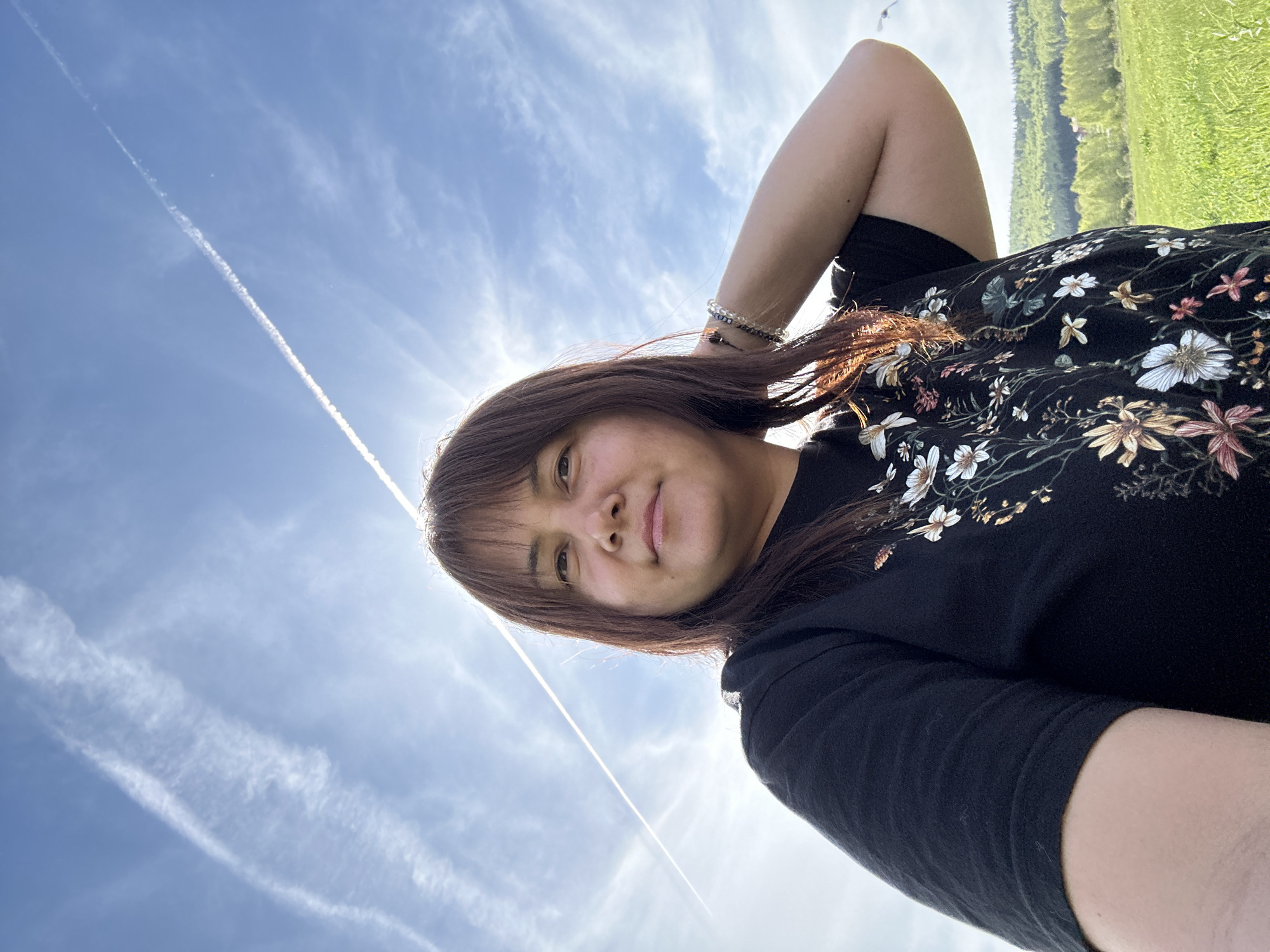 A person with brown hair and wearing a black floral shirt is lying down in a grassy field under a blue sky with white clouds and a contrail. The location is Unknown, Unknown.