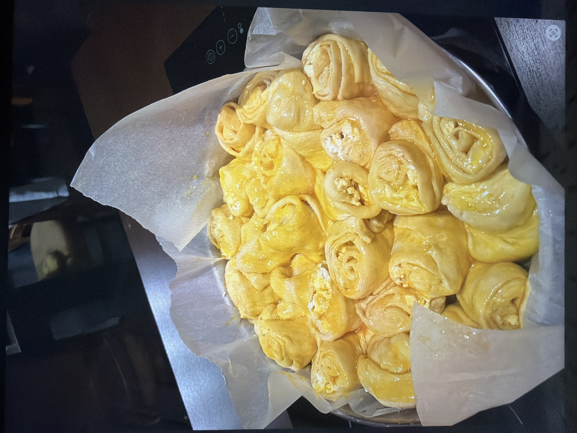 The image shows a close-up of a baking pan filled with unbaked, golden-yellow pastries arranged in a circular pattern. The pastries appear to be rolls, possibly filled with a light-colored substance such as cheese or custard. They are brushed with a glaze, making them appear glossy. The baking pan is lined with parchment paper. The scene appears to be indoors, likely in a kitchen setting, as indicated by the edge of a cooking appliance visible in the background. There is no indication that this image comes from the city of Burgas, Bulgaria.