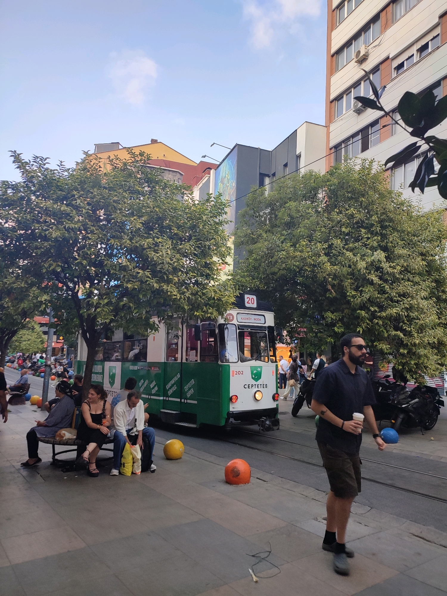 A street scene in Kadıköy, Turkey, features a green and white tram numbered 202 with "KADIKÖY-MODA" displayed on its front. The tram is on tracks running through a paved street lined with trees and buildings. Several people are visible, including a man with a beard and sunglasses walking with a cup in his hand, and a group of people sitting on a bench to the left. There are also yellow and orange bollards on the sidewalk and street. In the background, buildings of various architectural styles and heights, along with more trees and other people, contribute to the urban environment. The sky is partly cloudy.