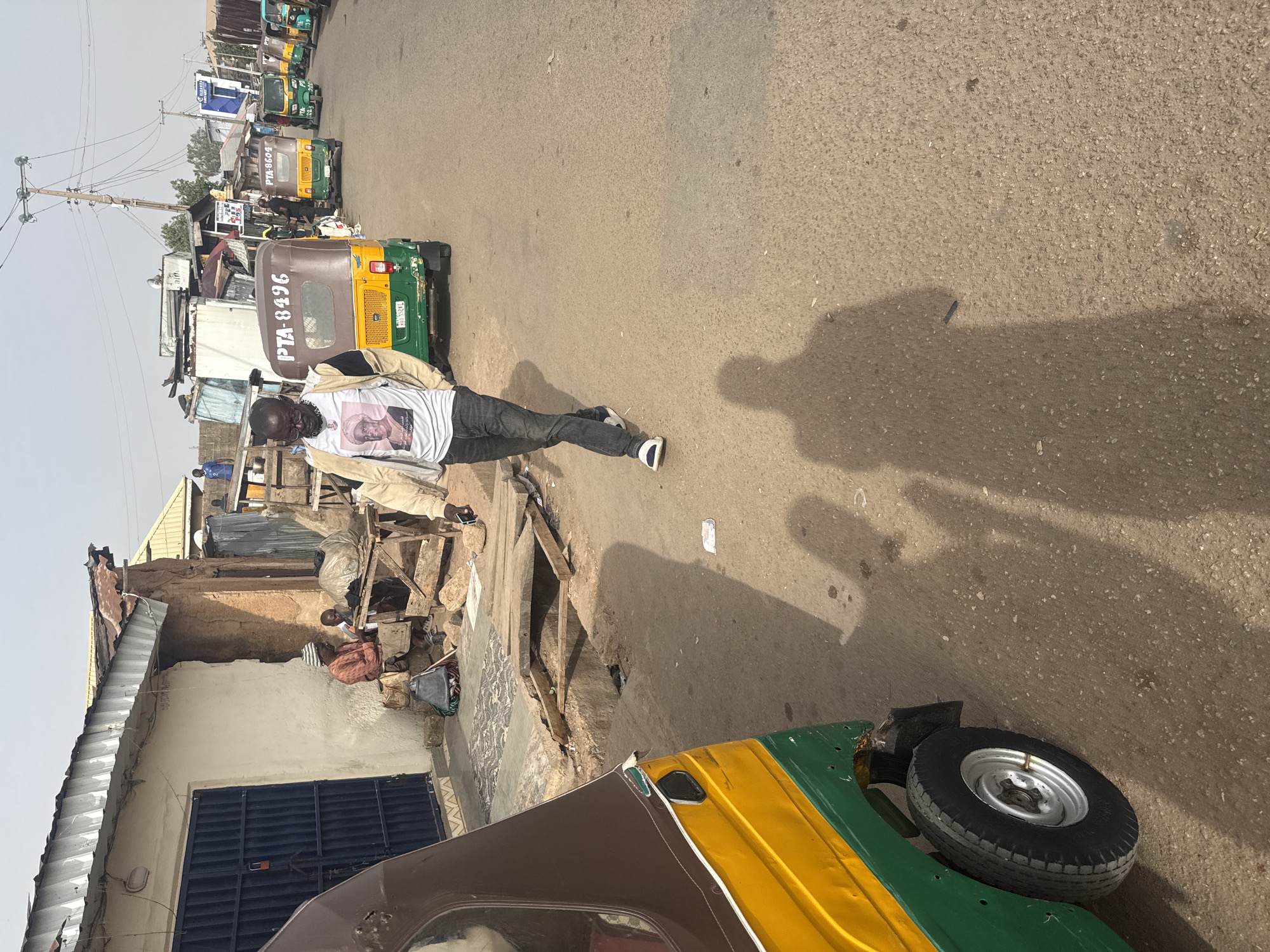 The image depicts a street scene in Jos, Nigeria, featuring a man walking down a paved road. He is wearing a white t-shirt with an image on it, dark pants, and sneakers. Several auto-rickshaws are parked along the side of the road. The rickshaws are primarily green, yellow, and brown, and one has the license plate "PTA-8496" visible. In the background, there are buildings of varying construction types, including a corrugated metal roof and a partially collapsed structure. A utility pole with wires is also visible. A man is seated by the side of a building.