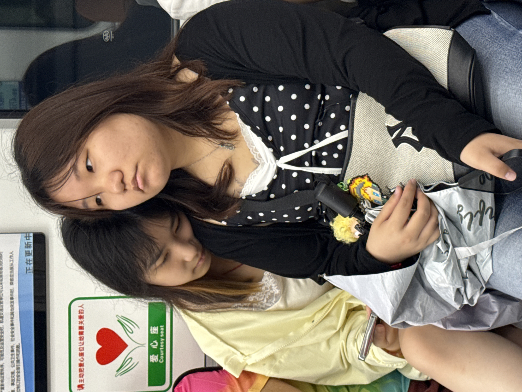 The image shows two young Asian women resting in what appears to be a public transport setting, possibly in Jiangjin, China. The woman in the foreground has long dark brown hair and is wearing a black long-sleeved top over a black and white polka dot garment with white lace trim. Her head is resting gently on the head or shoulder of the second woman, who has dark hair with lighter ends and is wearing a yellow long-sleeved top over a white lace-trimmed top, with her eyes closed, suggesting she is sleeping or deeply resting.
On the lap of the first woman, a black and white bag featuring the New York Yankees logo is visible, alongside a small, colorful plush charm. The second woman is holding a small silver object, possibly a phone or device, and a crumpled silver-grey bag with the stylized text "hfly" printed on it.
In the background, a blue and white sign contains partially visible Chinese text, including phrases like "...accidents and disasters, public health incidents, social security incidents and other emergencies, passengers should obey the staff...new safety operations. Updating..." A prominent green and white sign, common in public transport, features a red heart embraced by hands and the Chinese text "请主动把爱心座位让给需要关爱的人" (Please voluntarily give up the courtesy seat to those who need care) and "爱心座 Courtesy seat". The overall scene conveys a sense of peaceful rest during travel.