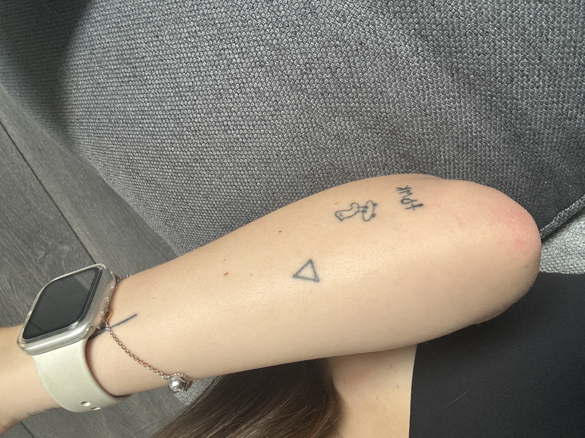 The image shows a close-up of a person's forearm resting on a gray fabric surface. The person is wearing an Apple Watch on their wrist with a light-colored band and a silver bracelet. There are three small tattoos on the forearm: a triangle, an outline of a person, and the word "FOUR" in a handwritten style. The background is out of focus and consists of gray fabric. There is also a hint of hair and black fabric. There is no information in the image to suggest a location in Huixquilucan de Degollado, Mexico.