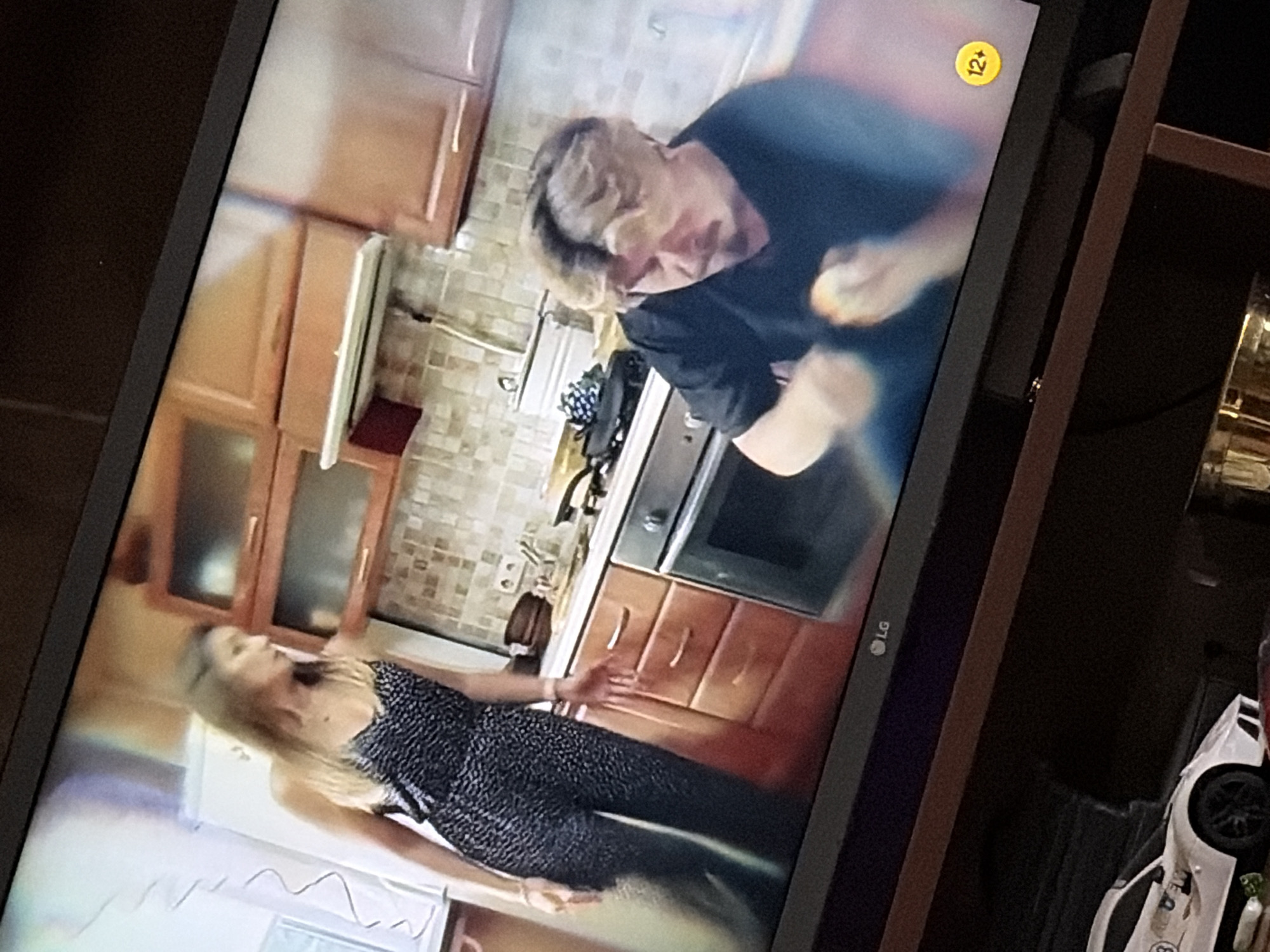A television screen displays a scene set in a kitchen. On the left, a woman with long blonde hair, wearing a black and white patterned jumpsuit, stands near a white refrigerator, looking to her right. In the center, a man with blonde hair and a mustache, wearing a dark t-shirt, is seated and appears to be peeling a piece of fruit. He is positioned in front of a kitchen counter with drawers and an oven. Above the counter, wooden cabinets are mounted on a tiled backsplash. A yellow circle with "12+" is visible on the lower right of the screen. Below the television, a white toy sports car is on a shelf, and a portion of a gold cylindrical object is also visible.