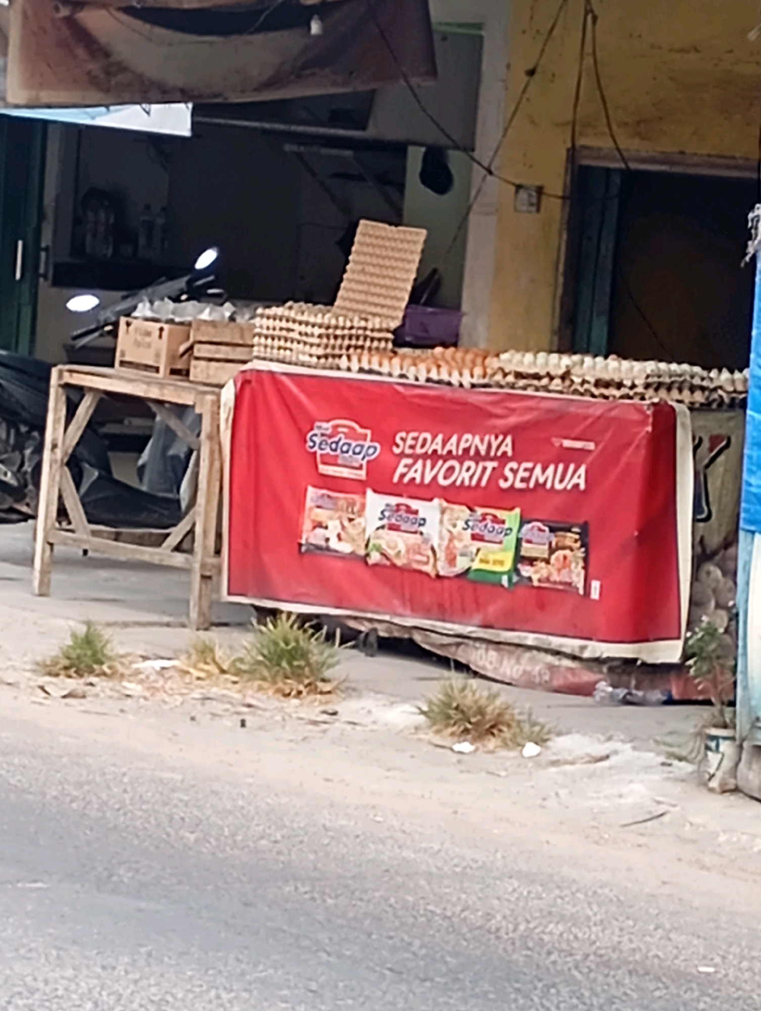 A street-level outdoor scene in Babelan, Indonesia, features a food stall with a prominent red banner displaying the text "Sedaapnya Favorit Semua" and images of various food products. The banner is stretched across the front of a display table laden with trays of eggs. To the left of the banner is a wooden stand holding cardboard boxes, and behind it, more trays of eggs are visible. The stall appears to be situated along a road, with a section of asphalt in the foreground and a building with yellow and blue accents in the background. Patches of dry grass and debris are scattered on the ground in front of the stall. The overall lighting suggests it is daytime, with natural light illuminating the scene. No individuals are clearly visible in the frame.