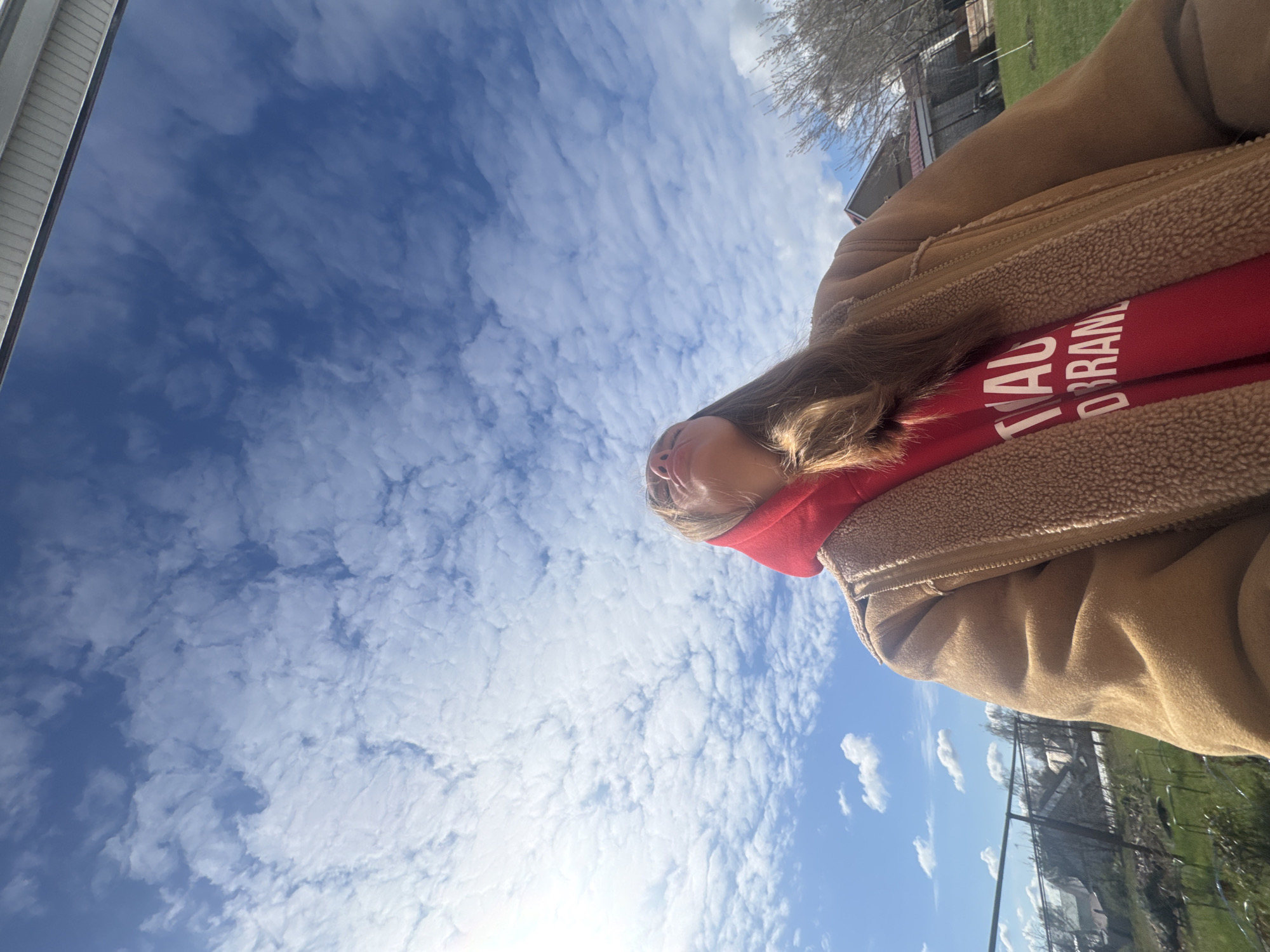 A low-angle outdoor shot depicts a person wearing a tan jacket with a fleece-lined collar and a red hooded sweatshirt with white lettering. The background is filled with a bright blue sky with scattered clouds. The person is positioned to the right of the frame, with a view of their face and the upper portion of their body. A glimpse of a building is on the upper left, and other buildings are on the upper right with the backdrop of green space. A wire fence also borders the area of the shot in the upper right. The location details (Bol'shoye Kozino, Russia) are not visibly verifiable within the image.