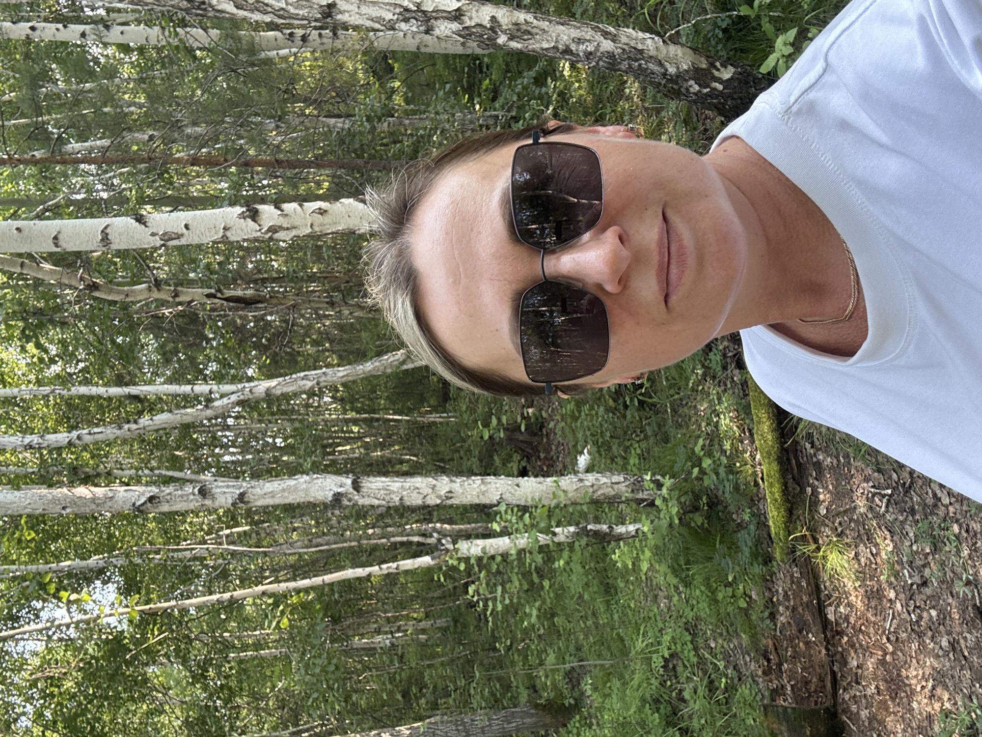 The image is a selfie of a woman with blonde hair, wearing large dark sunglasses and a white t-shirt. She is standing in a forest with many birch trees. The ground is covered in fallen leaves and moss. The woman is smiling slightly. The location is identified as the city of Bratsk, Russia, suggesting this is an outdoor scene within or near the city.
