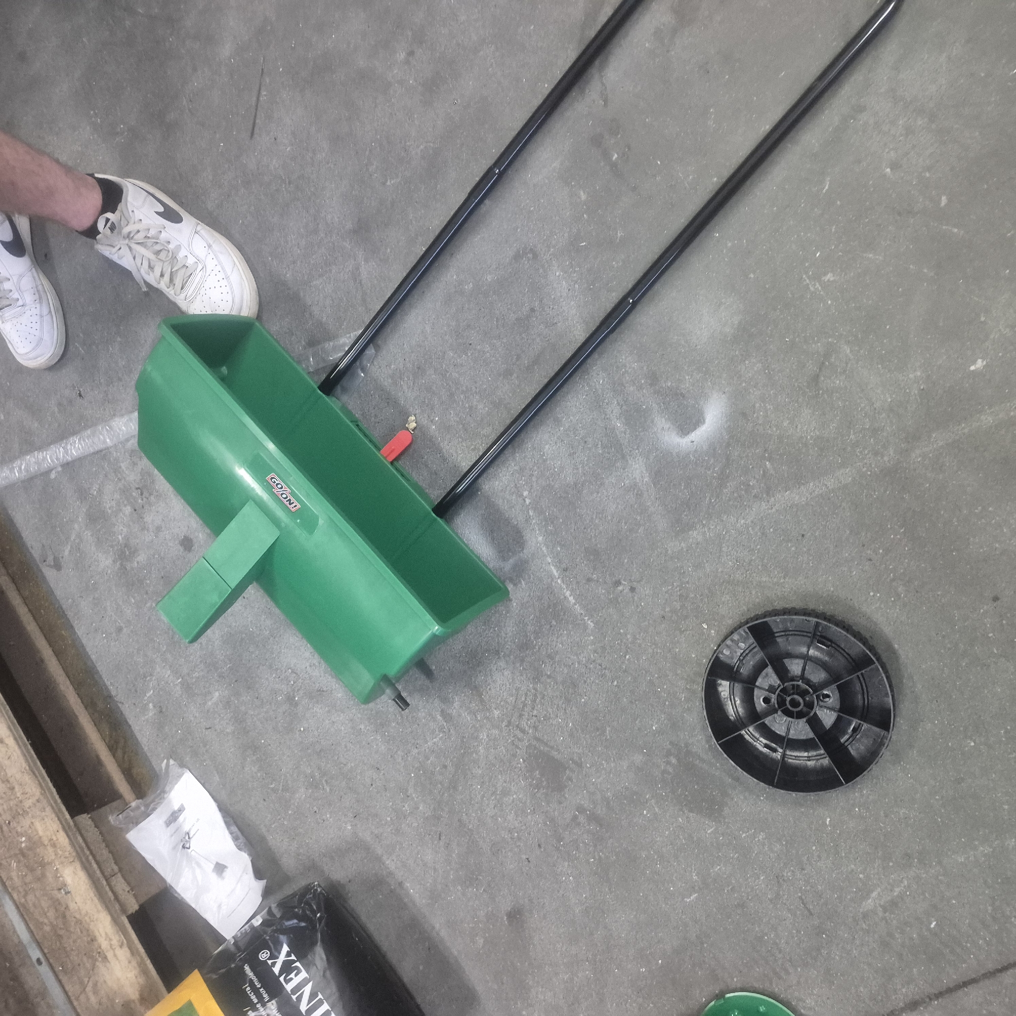 The image shows a green seed spreader lying on a concrete floor, possibly in an industrial setting, as suggested by a wooden pallet in the background and a bag labeled "LINEX". Two black metal poles extend from the seed spreader, suggesting it is designed for walking or pushing. To the left of the spreader is a black plastic wheel with a spoke design. In the background, a person's feet wearing white Nike sneakers are visible. The overall scene is consistent with a storage area or workshop. The image does not provide any information about the city of Nowa Wieś Książęca, Poland.