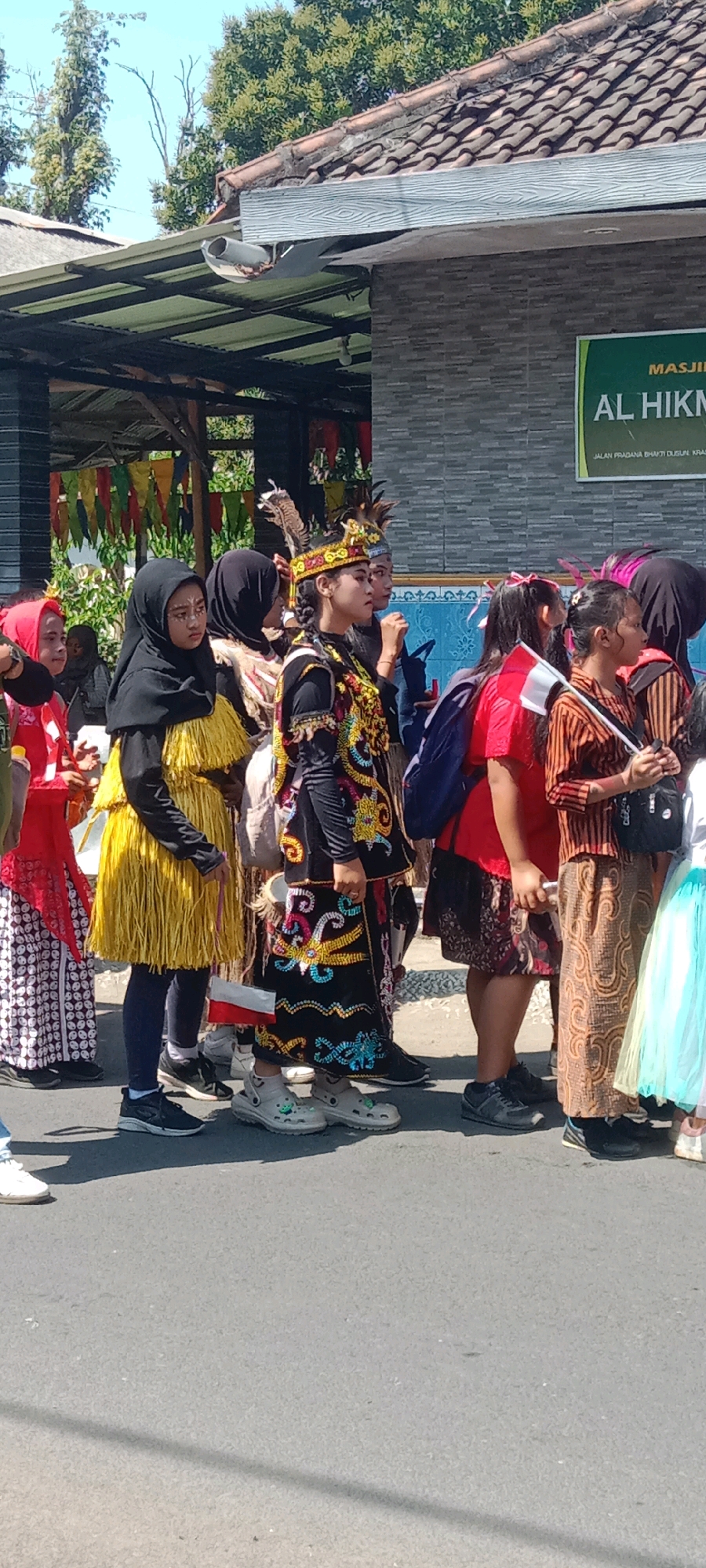 A group of people, primarily women and girls, are standing in what appears to be a procession or parade on a paved street. Several individuals are wearing traditional clothing, including ornate headdresses, colorful embroidered vests, and patterned skirts. Some are wearing hijabs. A sign with green lettering reads "MASJID AL HIKMA" and below it, "JALAN PRADANA BHAKTI DUSUN KRAS." This indicates the location is Masjid Al Hikma, situated on Jalan Pradana Bhakti, in the village of Kras, Indonesia. The background shows a brick building with a tiled roof and some trees. The lighting suggests it is daytime with bright sunlight. The people are facing forward and seem to be waiting or moving in a line.