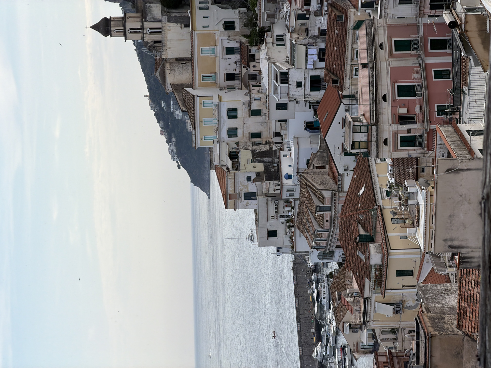 A high-angle, vertical shot shows an Italian coastal town built on a steep slope. The foreground features a jumble of closely packed buildings, mostly white or pastel colored, with orange tile roofs. Many windows are visible, often with green shutters. Clothes hang on some lines. The buildings appear stacked upon each other, typical of hillside towns.
In the middle ground, a body of water stretches out to the left. A stone pier runs along the edge of the water, filled with boats. Across the water, a mountain covered in vegetation is visible. A tall, thin tower with a pointed roof stands out against the mountain.
The sky is visible on the left of the frame, light blue and slightly cloudy. The overall scene presents a typical landscape of the Amalfi Coast.