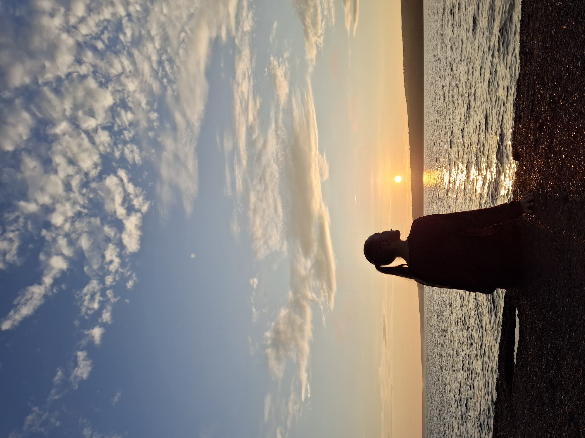 A silhouette of a young woman with her hair in a ponytail is seated on a pebbled shoreline, facing a body of water. The sun is setting, casting a golden glow on the water and the sky. The woman is wearing a dark, long-sleeved shirt. The water is calm, with gentle ripples reflecting the sunlight. In the distance, a treed shoreline is visible, and the sky is filled with scattered clouds. The scene appears to be a serene natural setting, possibly a lake or riverbank, in the city of Chaikovsky, Russia, given the prominence of the Kama River and its surrounding landscape in the area.