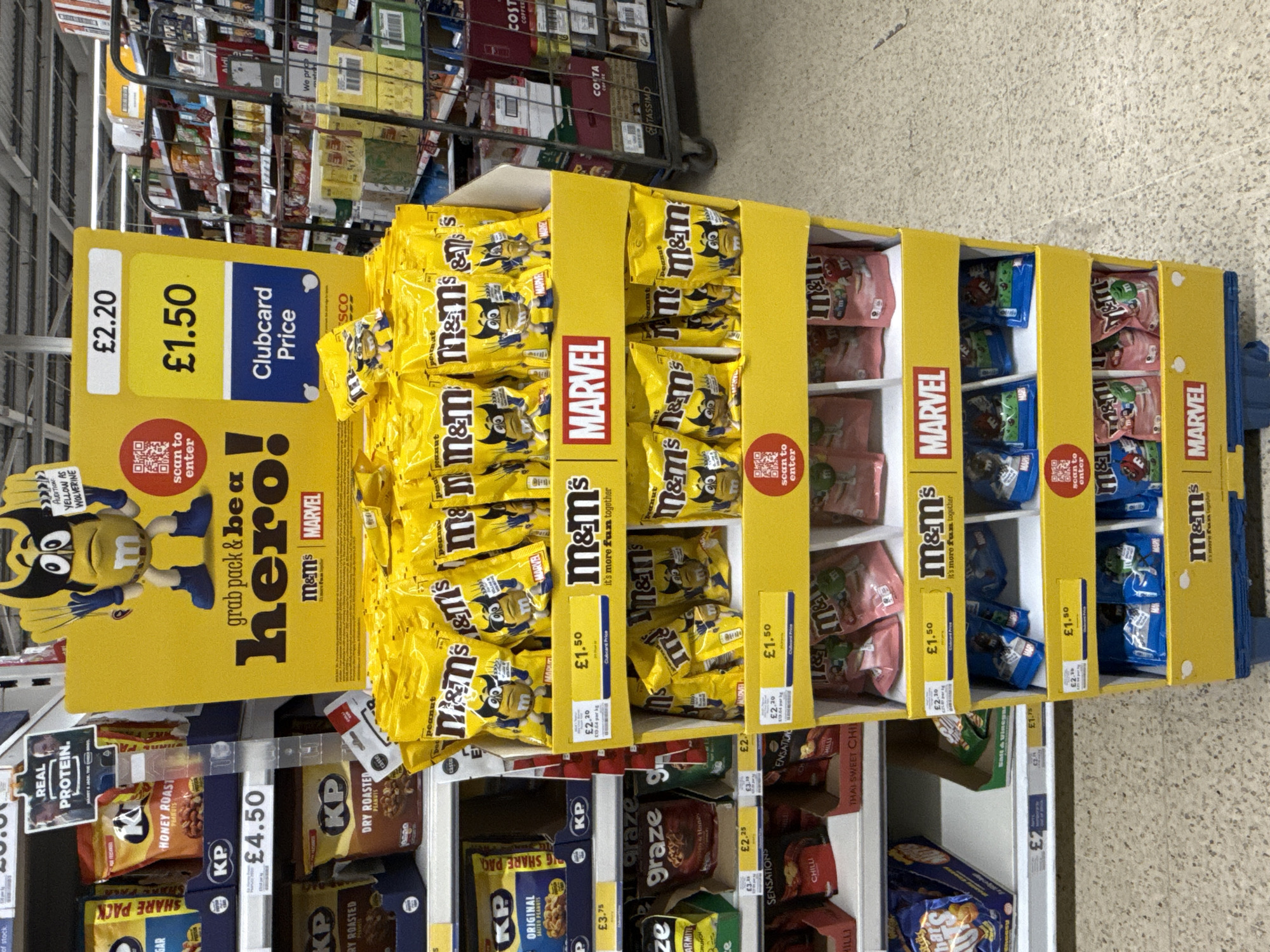 This image shows a promotional display for M&M's candies situated in a supermarket aisle, likely in Great Yarmouth, United Kingdom, given the currency and common UK brands. The display is predominantly yellow and features M&M's in a Marvel Comics collaboration, specifically highlighting the yellow M&M character dressed as Wolverine.
The central part of the image focuses on a multi-tiered display stand, heavily stocked with bags of yellow Peanut M&M's featuring the Wolverine character. Pink and blue varieties of M&M's are also visible on different shelves of the display. A prominent sign on the left side advertises a price of "£2.20" with a "Clubcard Price" of "£1.50" for the M&M's. Individual price tags throughout the display reiterate this offer.
Various promotional texts are visible on the display, including "grab pack & be a hero!", "m&m's MARVEL", "AUDITION YELLOW AS WOLVERINE", and "it's more fun together". Several QR codes with the prompt "scan to enter" suggest an interactive element or competition.
Below and to the sides of the M&M's display, standard supermarket shelving holds other snack products. These include multiple varieties of KP-branded peanuts, such as "Honey Roast Peanuts," "Dry Roasted Peanuts," and "Original Salted Peanuts," with some packs priced at "£4.50" and "£3.75." Brands like Graze snacks and Sensations crisps are also visible. In the background, further down the aisle, shelves are stocked with various packaged goods, including coffee products like Costa and Tassimo, and a metal shopping cart is present. The scene depicts a typical indoor retail environment.