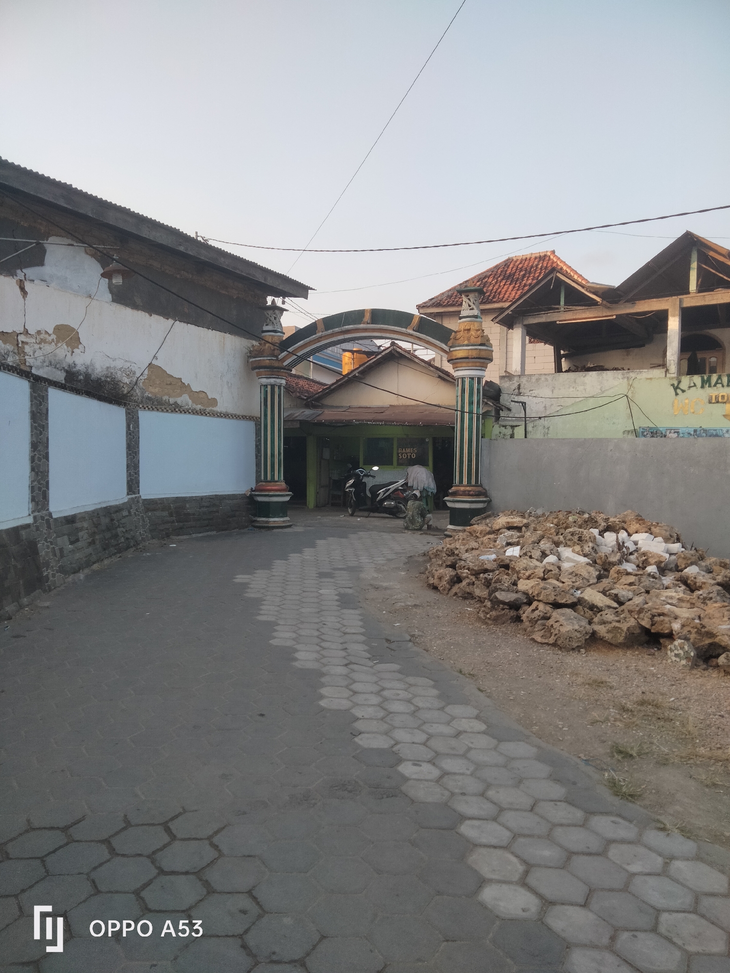The image captures an outdoor scene in what appears to be a residential or commercial area in Badung, Indonesia. A paved walkway with a hexagonal pattern leads towards an arched entryway. The entryway features decorative pillars with green and gold accents. Beyond the arch, a building with a green facade and signage that reads "RAMES SOTO" is visible, with a motorcycle parked to its left. To the right of the walkway, a pile of rough, light-colored stones is stacked. The architecture suggests a traditional Indonesian style, with visible signs of wear and tear on some of the surrounding buildings, including cracked plaster on a white wall on the left. The sky is a clear, pale blue, indicating daylight. The overall impression is of a humble, everyday setting within a city.
