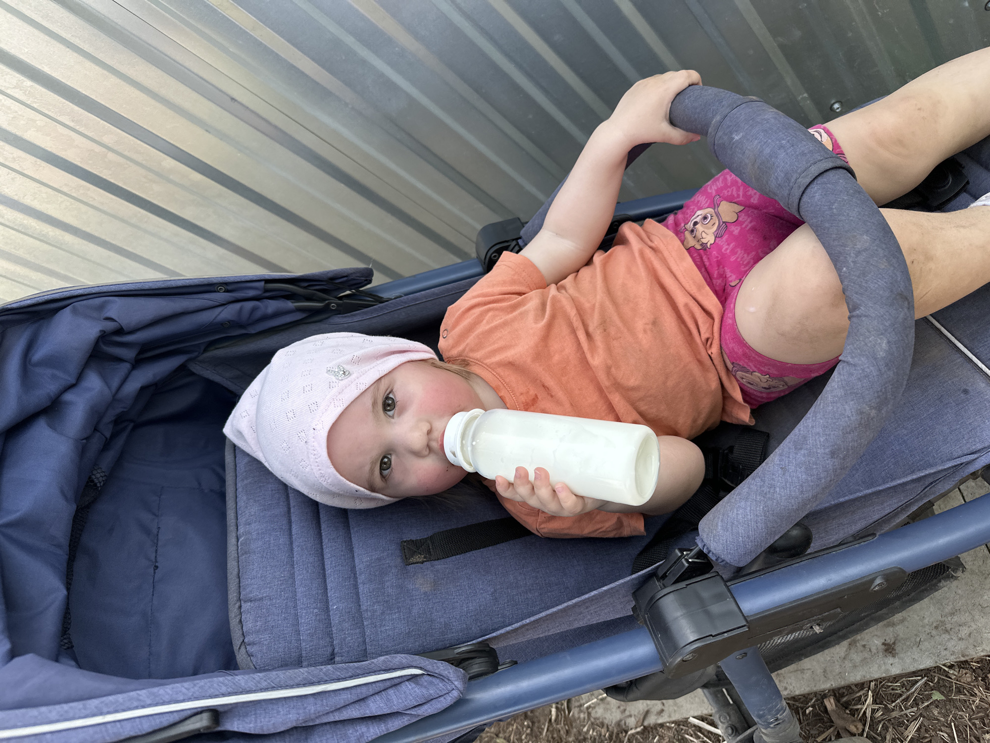 A toddler lies in a blue stroller, angled to the left, holding a white baby bottle to their mouth. The child wears a light pink bonnet, an orange shirt, and pink shorts with a cartoon character printed on them. The stroller is parked on a grey concrete surface adjacent to a corrugated metal wall. The setting is in the city of Chaikovsky, Russia.