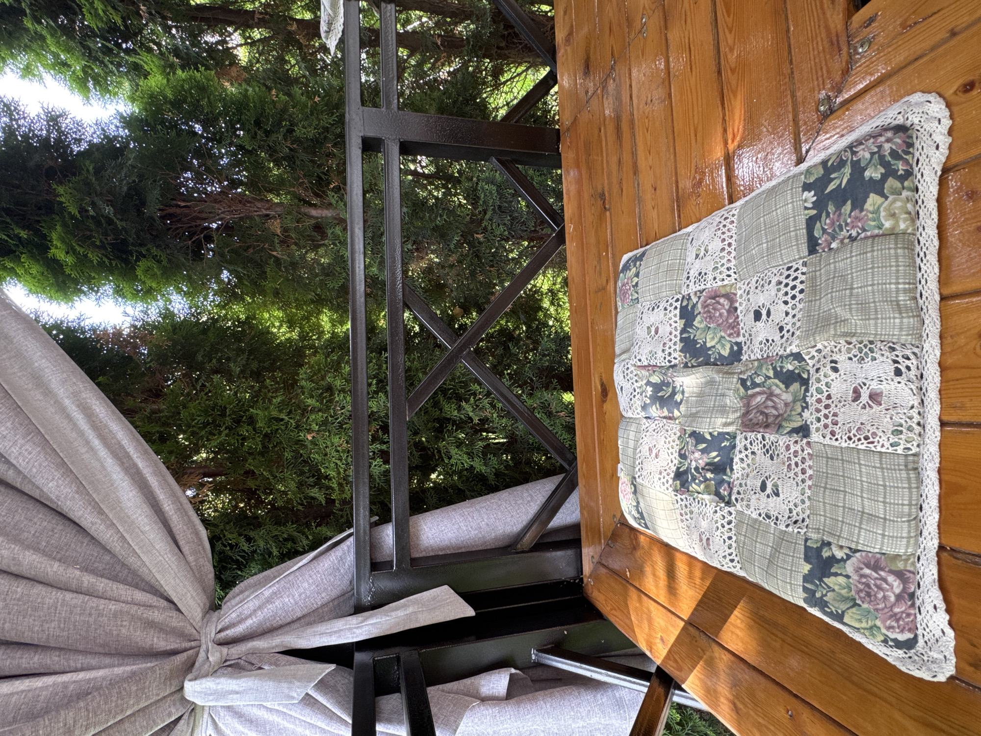 A close-up, eye-level shot shows a quilted cushion on a wooden table outdoors. The cushion, placed on the right side of the table, is made of squares of floral fabric, plaid fabric, and crochet lace, with a crocheted edge. The table is made of polished wooden planks. In the background, trees are visible, with a dark metal railing in front of them. A light gray curtain is tied back on the left side of the image, obscuring part of the background. The setting appears to be a covered outdoor seating area, possibly a balcony or veranda, in Sary-Oy, Kyrgyzstan.