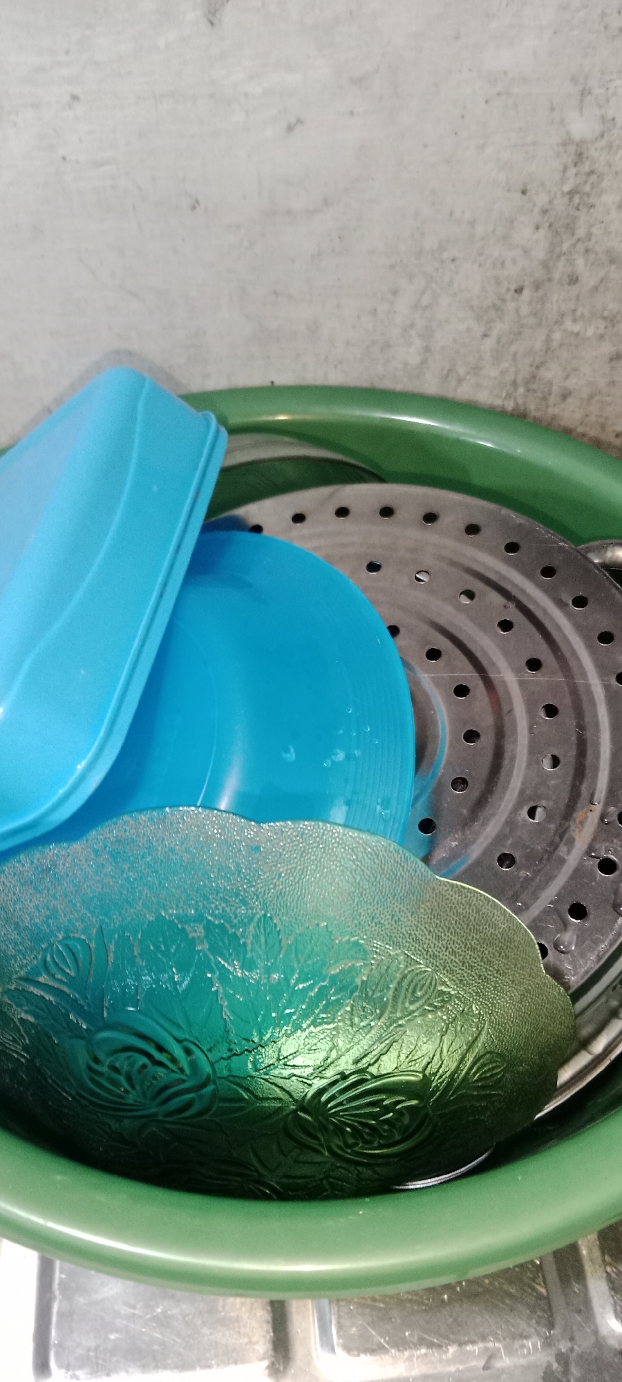 The image shows a close-up of several kitchen items stacked inside a green plastic basin. The basin contains a metal steamer insert with numerous holes, a blue plastic plate, and a blue plastic lid. Below these items, a green glass bowl with an embossed floral pattern is visible. The background appears to be a textured wall. The scene is indoors, likely in a kitchen setting in Subang, Indonesia, based on the provided information. There are no people or discernible actions taking place, the focus is on the arrangement of these household objects.