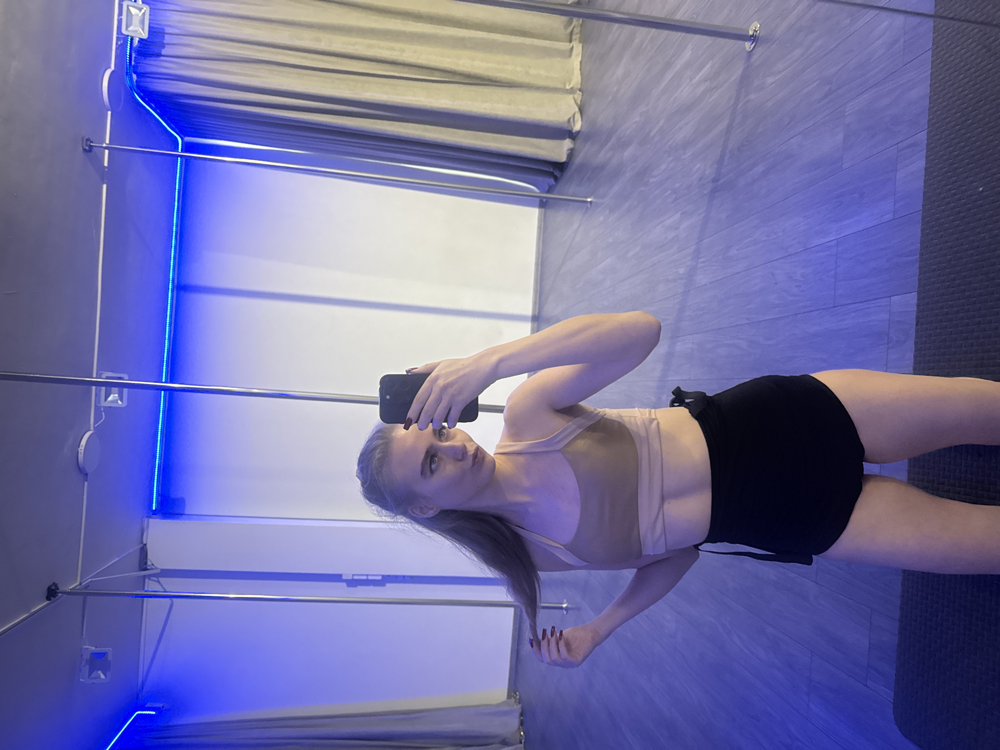 A female subject is depicted taking a mirror selfie within an indoor studio, location Unknown, Unknown. She has light-colored hair, fair skin, and is dressed in a light tan sports bra and black shorts, with red nail polish visible on her fingernails. Her right hand holds a dark smartphone, pointed towards her reflection, while her left hand touches a section of her hair. The studio environment features multiple chrome vertical poles extending from floor to ceiling, large wall mirrors, and blue LED strip lighting positioned along the upper perimeter, casting a blue hue. The floor is light grey wood-look laminate, with a dark textured mat in the immediate foreground beneath the subject's feet. Heavy beige curtains are visible in the background, both directly and as reflections. The ceiling is dark grey with mounted square light fixtures.