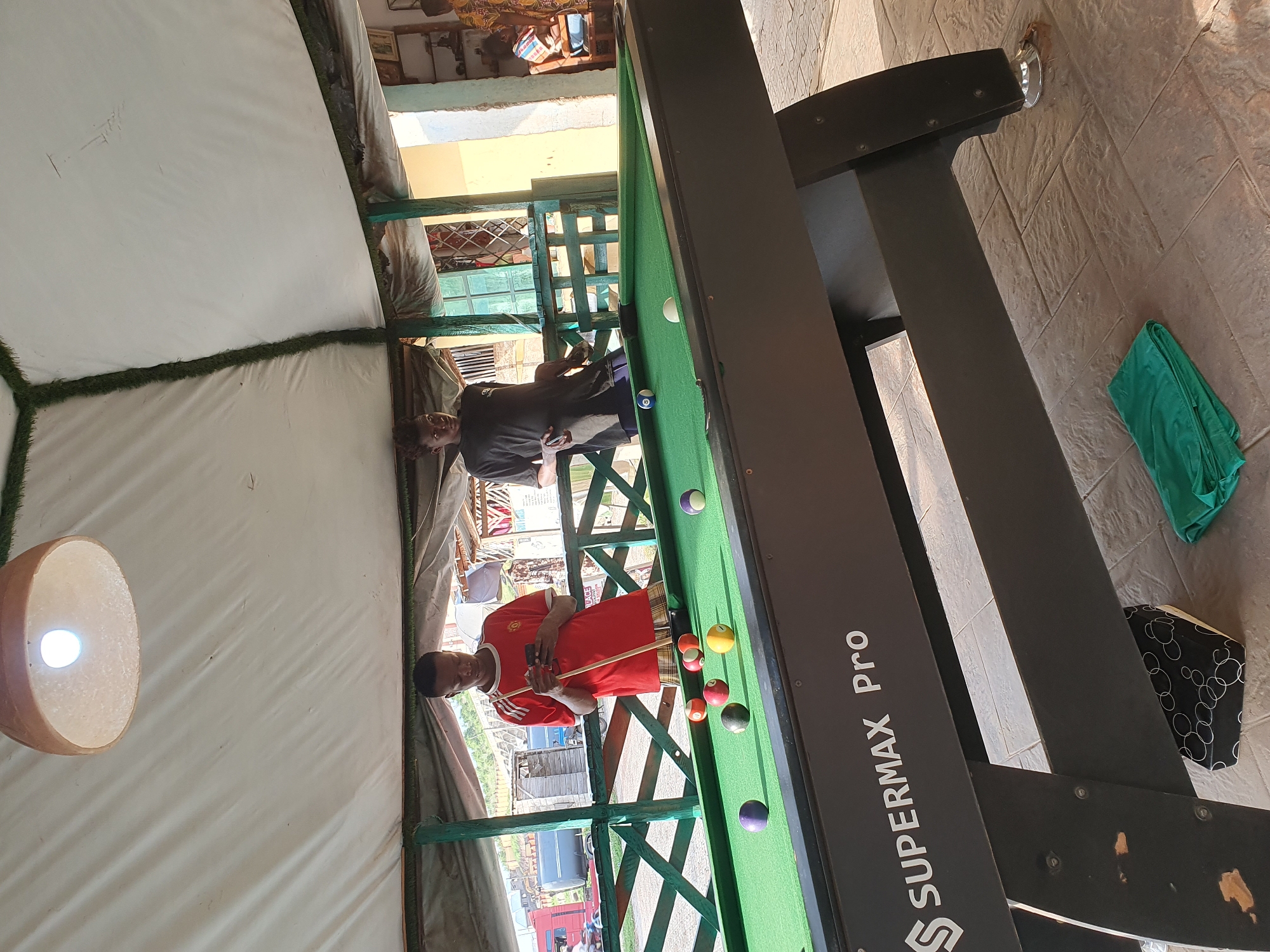 The image depicts an outdoor covered area, likely a bar or recreational space, located in Lokoja, Nigeria. Two young men are visible. One, wearing a red t-shirt and patterned shorts, stands by a pool table, holding a pool cue and looking at his phone. The other, in a dark t-shirt and dark pants, leans against a railing near the pool table, looking towards the camera. The pool table itself is green with several colored balls scattered across its surface. A light fixture hangs from the white tent-like ceiling above. In the background, there are glimpses of buildings, a road with vehicles, and some market stalls. The floor is paved with textured tiles. A folded green cloth lies on the ground near the base of the pool table. The overall impression is of a casual, possibly public, gathering place.