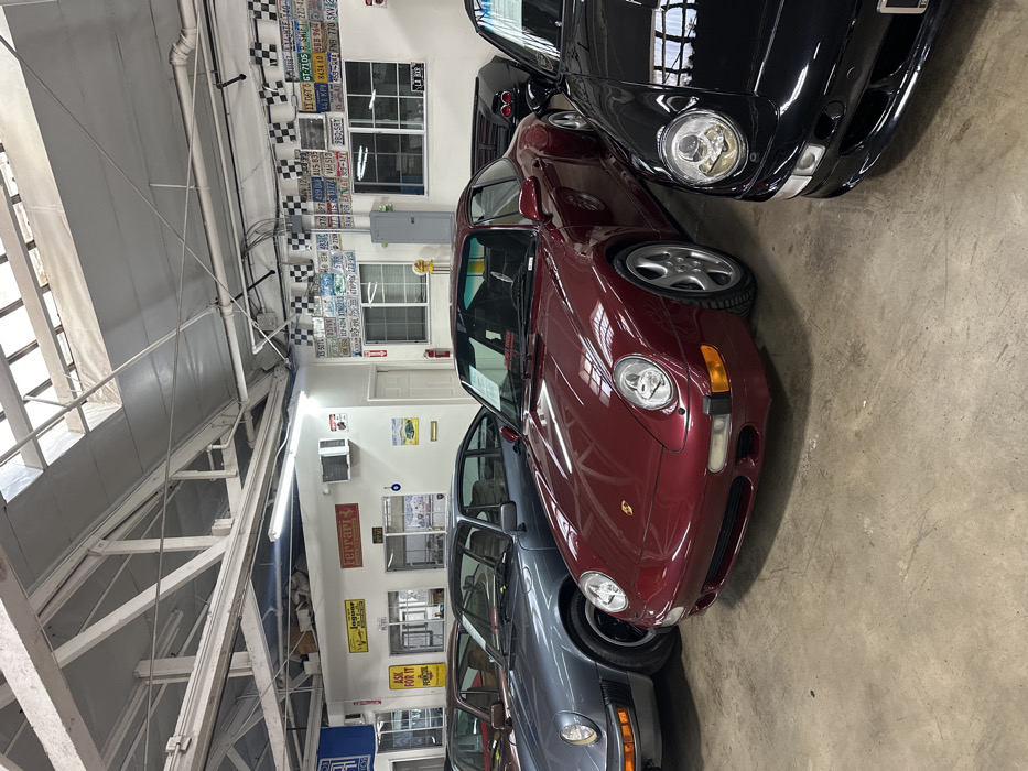 The image showcases four Porsche 911s parked closely together in what appears to be a showroom or garage. The cars vary in color, including black, red, and gray. The space is decorated with automotive memorabilia like license plates and signs. A “Ferrari” sign, along with others reading “Ask For It,” and “Labor” can be seen. There are also license plates that appear to read “11 06T GT-7105 PSHIFT 2204 48434 1088 964”. The interior structure has white beams and windows that let in natural light.