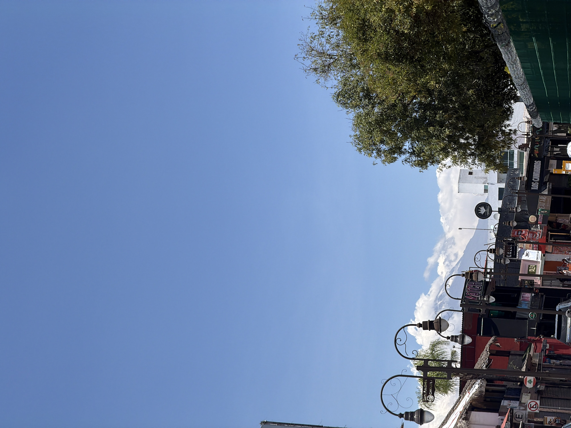 The image shows a view of San Andrés Cholula, Mexico, looking upwards. The sky is mostly clear blue. In the top right corner, there is a tree with dense green foliage. Below the tree, there is a street scene with various buildings, signs, and streetlights. The buildings are a mix of white and dark colors. Several streetlights with ornate curved arms are visible. The street is lined with signs and various advertisements. A mountain is visible in the background, partially obscured by clouds.