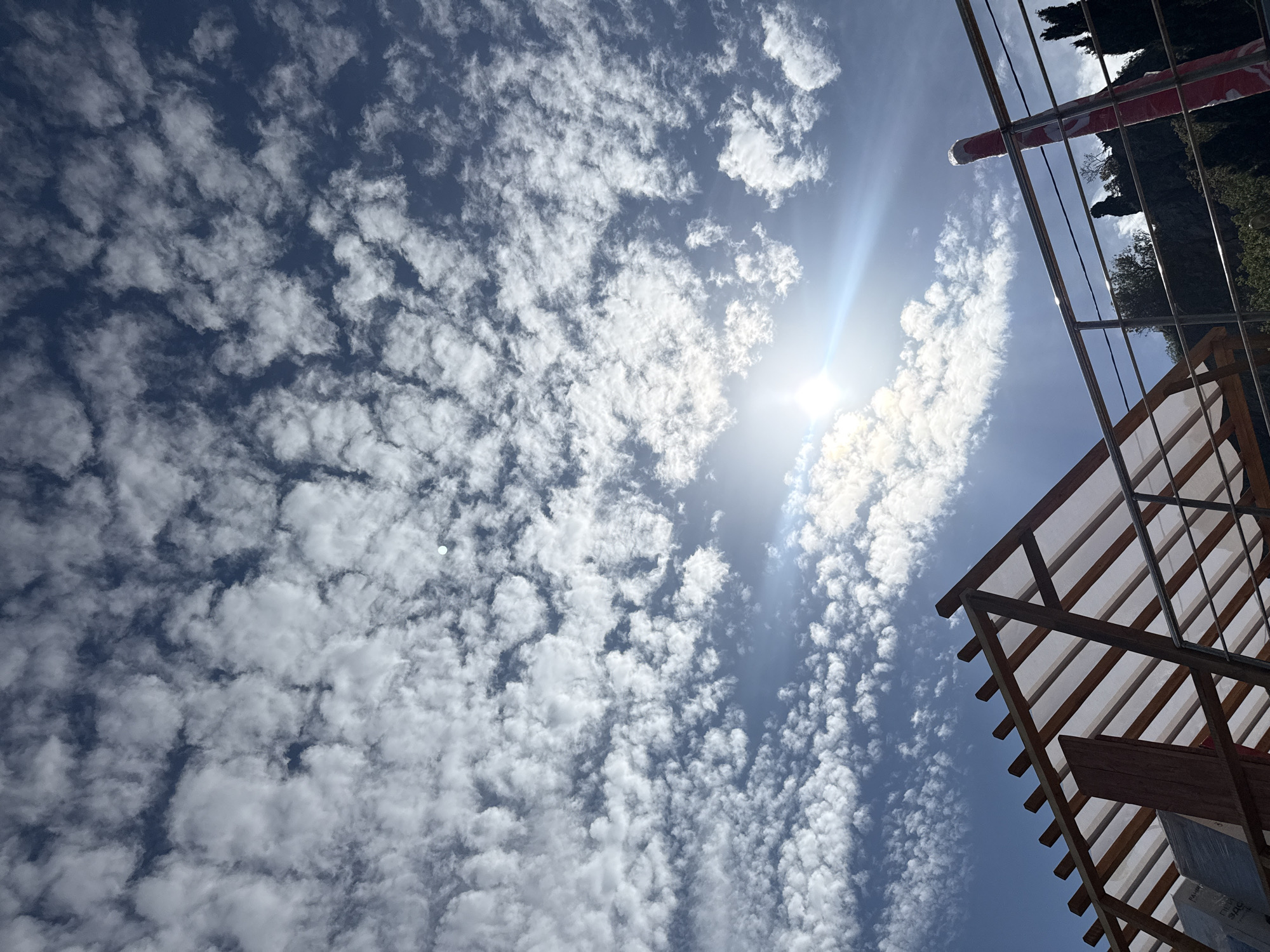 The image captures a bright sunny day with a sky filled with numerous small, fluffy white clouds, possibly altocumulus or cirrocumulus. The sun is high in the sky, creating a glare. A portion of a wooden structure, possibly a pergola or outdoor shelter, is visible in the lower right corner of the image, with some metal elements present. Some trees and a red and white striped object can be seen at the top right, hinting at a location near a building and a flag. The environment suggests an outdoor space, possibly a garden or patio. The city is Unknown, Unknown. There are no people visible in the image.