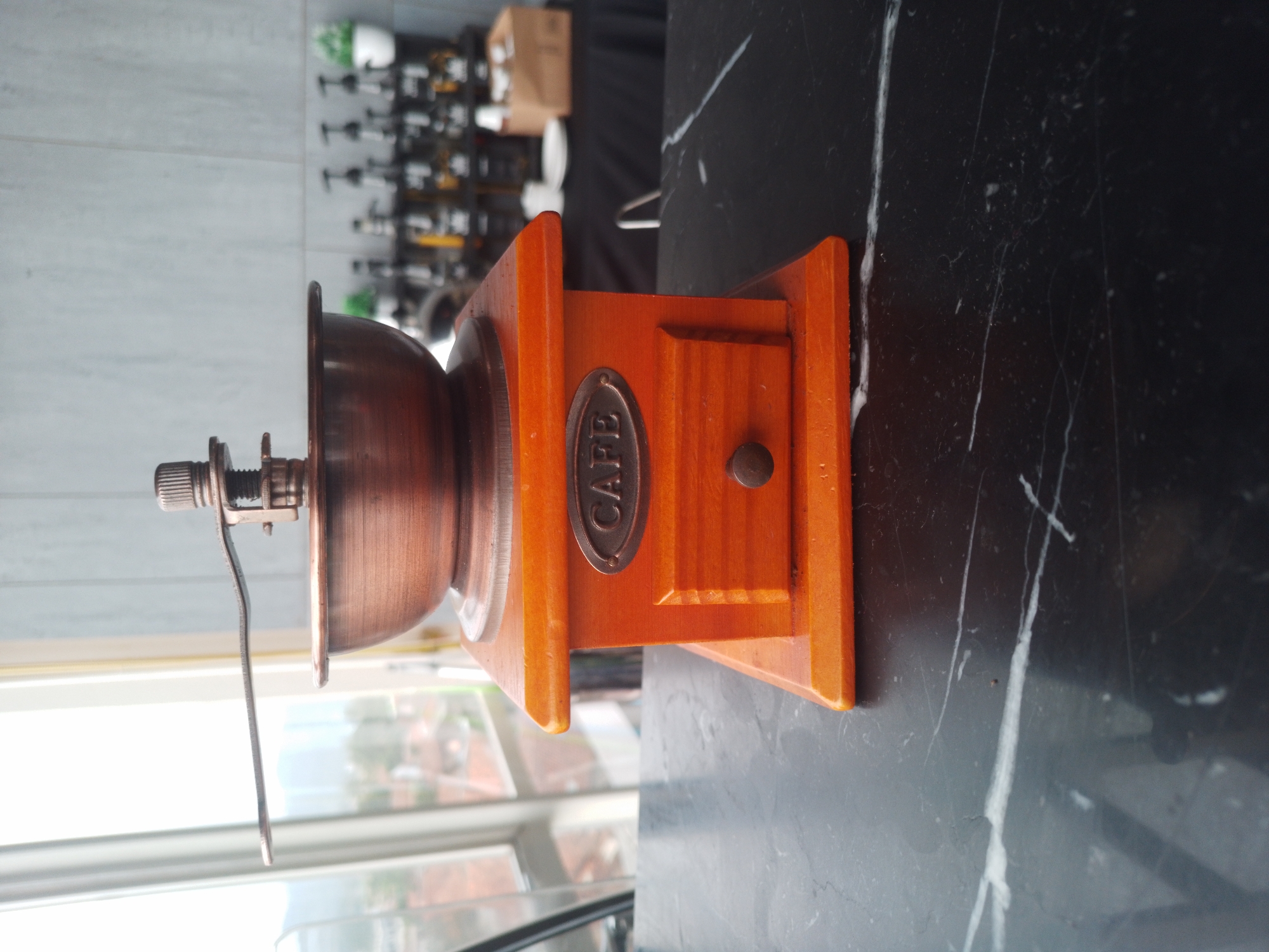 A vintage-style manual coffee grinder with a copper bowl and a wooden base sits on a black marble countertop. The grinder has a wooden drawer with a small knob. The word "CAFE" is embossed on a metal plate on the front of the wooden base. In the background, a blurred scene shows shelves with bottles, possibly syrup dispensers, and some greenery, suggesting a cafe or coffee shop setting. A window with a view of buildings and trees is also visible in the background. The image is taken from a medium-low angle.