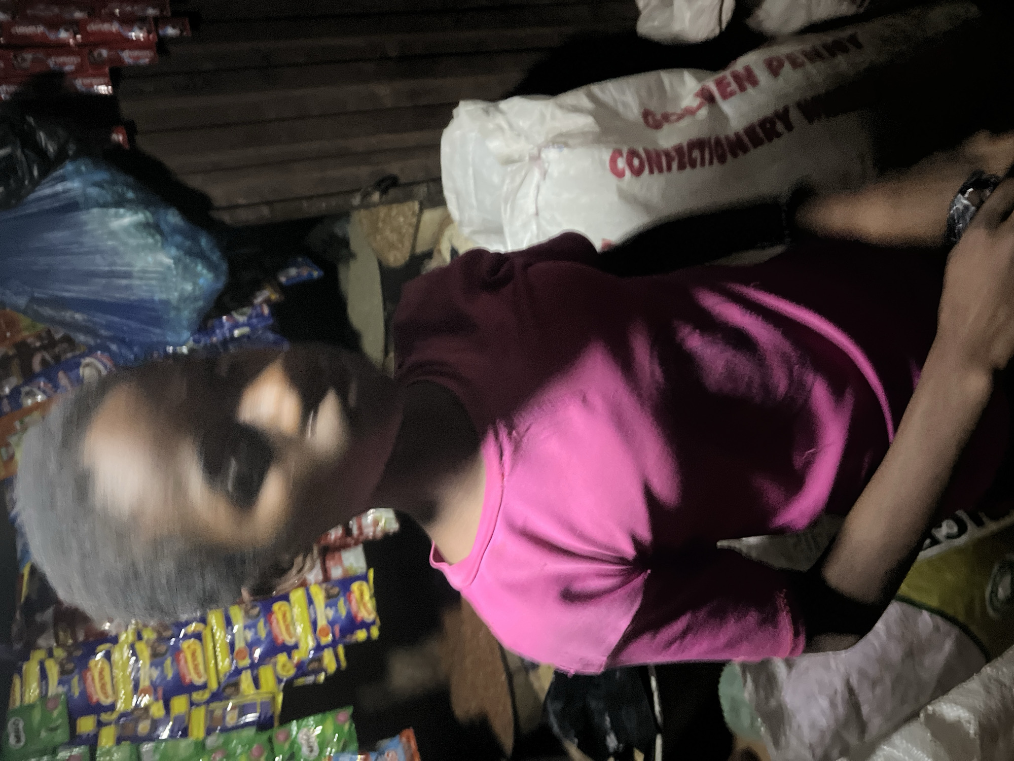 A person in a pink shirt is lying among stacks of confectionary items, next to a large bag labeled "Golden Penny Confectionery." This scene is occurring in an area where goods are being sold. The specific location within Abuja, Nigeria, cannot be determined from the image.