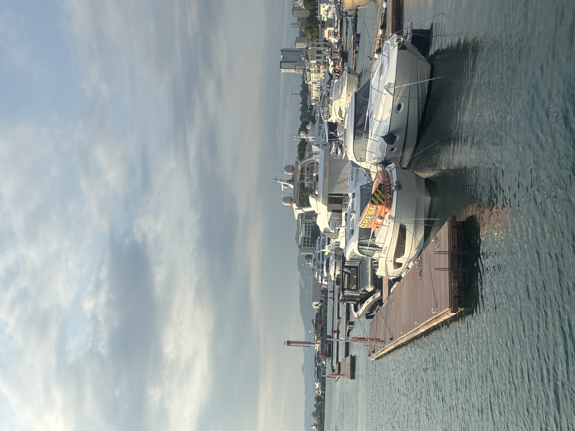 The image depicts a marina in Sochi, Russia, with several yachts docked. The water is calm and reflects the overcast sky. In the foreground, a wooden pier extends into the water, with two white yachts moored alongside. One of the yachts has an orange life preserver visible. In the background, a cityscape with buildings and a tall, red and white striped tower are visible. The sky is filled with clouds, suggesting either early morning or late afternoon. There are no people visible in the image.