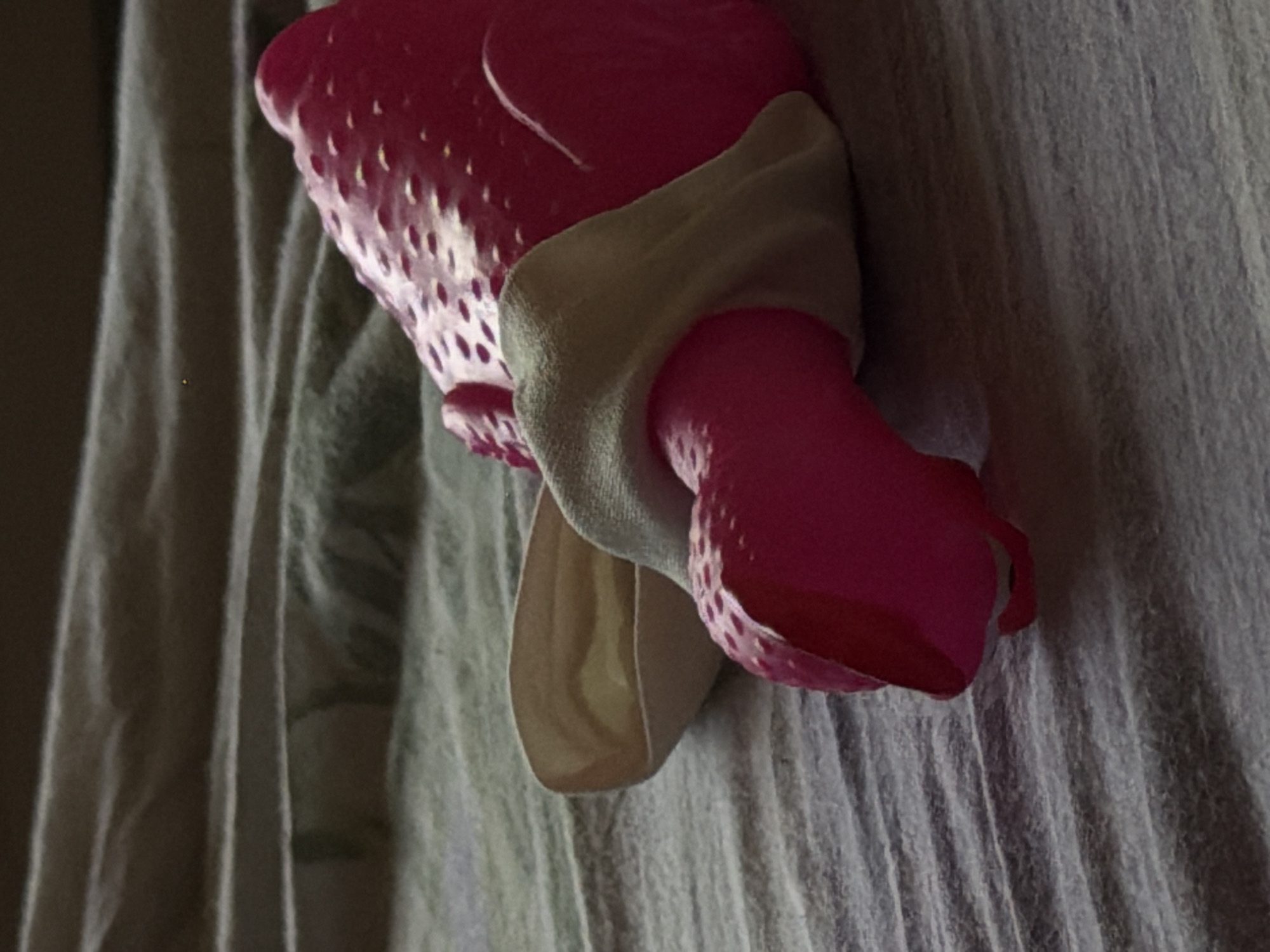 A close-up, low-angle, eye-level shot shows a pink, textured, rubber toy lying on a grey, ribbed surface, possibly a blanket or bedsheet. The toy appears to be a stylized animal, with a rounded body and raised bumps across its surface. A light-colored fabric is draped over the toy's lower section. The lighting is dim, casting shadows and highlighting the textures of both the toy and the surface it rests on. The background is out of focus, suggesting the image was taken indoors, with no visible signs of the city of Bobrovo, Russia.