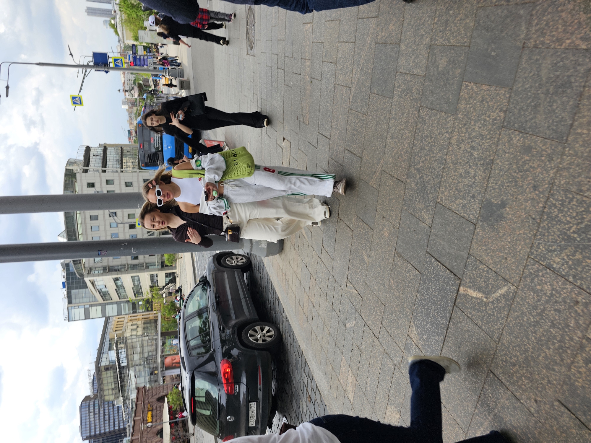 The image captures a group of four women on a city street, likely in Moscow, Russia, based on the presence of a vehicle license plate with the country code. The women appear to be walking together. One woman wears black pants, a black top, and carries a black bag. Another wears white pants with green stripes and a white top. A third woman wears white pants and a black top. The fourth woman wears all black. The background shows a typical urban landscape, including buildings, street signs, and a bus. A dark-colored car is parked adjacent to the sidewalk where the women are walking. The scene is set during daylight hours under a partially cloudy sky.