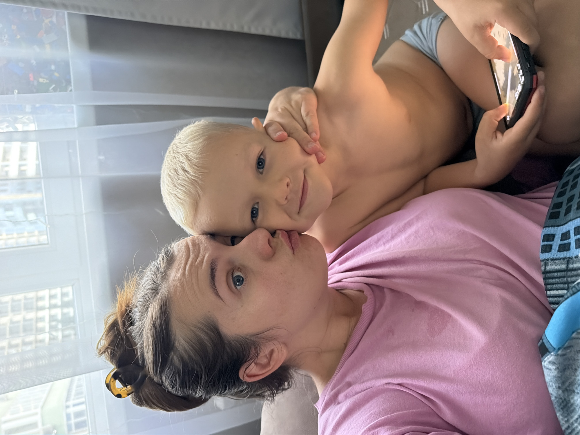 The image depicts a tender and playful moment between a woman and a young child indoors, likely in Sochi, Russia. The photo is taken at an angle, showing them in close proximity, possibly lying down together.
The woman, with light brown hair pulled back with a tortoiseshell claw clip, wears a pink t-shirt. Her blue eyes are wide, and her lips are slightly puckered in a gentle or affectionate expression as she looks towards the child.
A young boy, with short blonde hair and bright blue eyes, is positioned on top of or closely beside her. He is shirtless, wearing grey underwear or shorts, and looks directly at the camera with a faint, sweet smile. With both hands, he holds a black smartphone with red accents, its illuminated screen suggesting he is engaged with content on the device.
They appear to be on a soft surface, possibly a sofa or bed, indicated by grey and blue patterned fabric. Bright daylight streams in from a large window behind them, diffused by white horizontal blinds. Through the blinds, the outlines of urban buildings are visible, consistent with a city setting like Sochi. The overall mood conveyed is one of warmth, intimacy, and everyday family connection. No legible text is visible in the image.