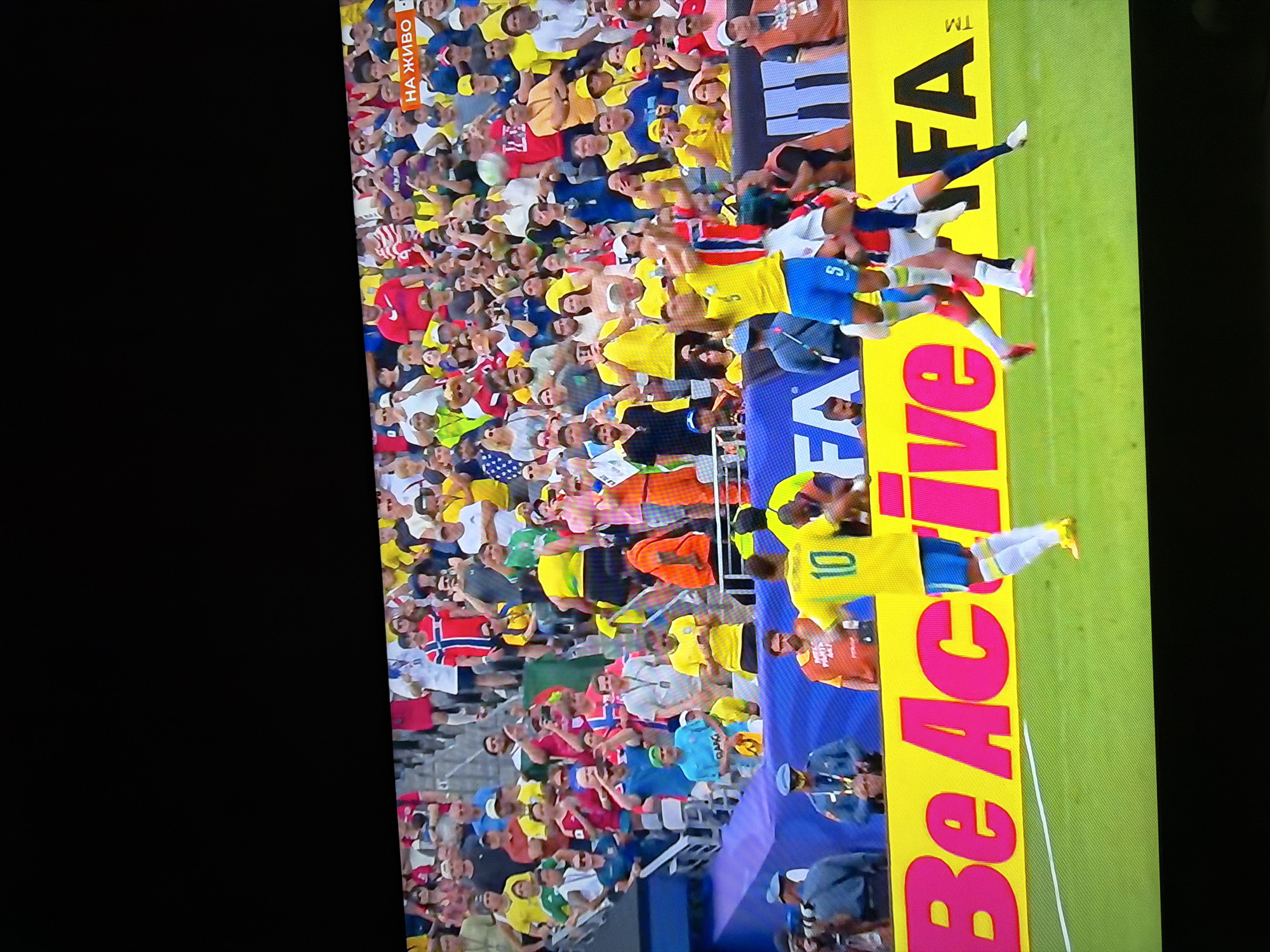 The image captures a soccer match, likely a broadcast on a television screen, as indicated by the screen's black border and the glare. A crowd of spectators, many wearing team colors and national flags, fills the stadium seating. The field itself is green, with a bright yellow advertising board running along the edge. On the board, the words "Be Active" are prominently displayed in large, pink text, followed by "FIFA". In the foreground, two soccer players in uniform are visible. One player, wearing a yellow jersey with the number 10 and blue shorts, is in motion. Another player in a red and white striped jersey is also in the frame, appearing to be in a defensive or celebratory pose. The overall scene depicts a dynamic moment during a sporting event, with the audience actively engaged. The context of Ognianovo, Bulgaria, is not directly evident within the visual elements of the image itself.
