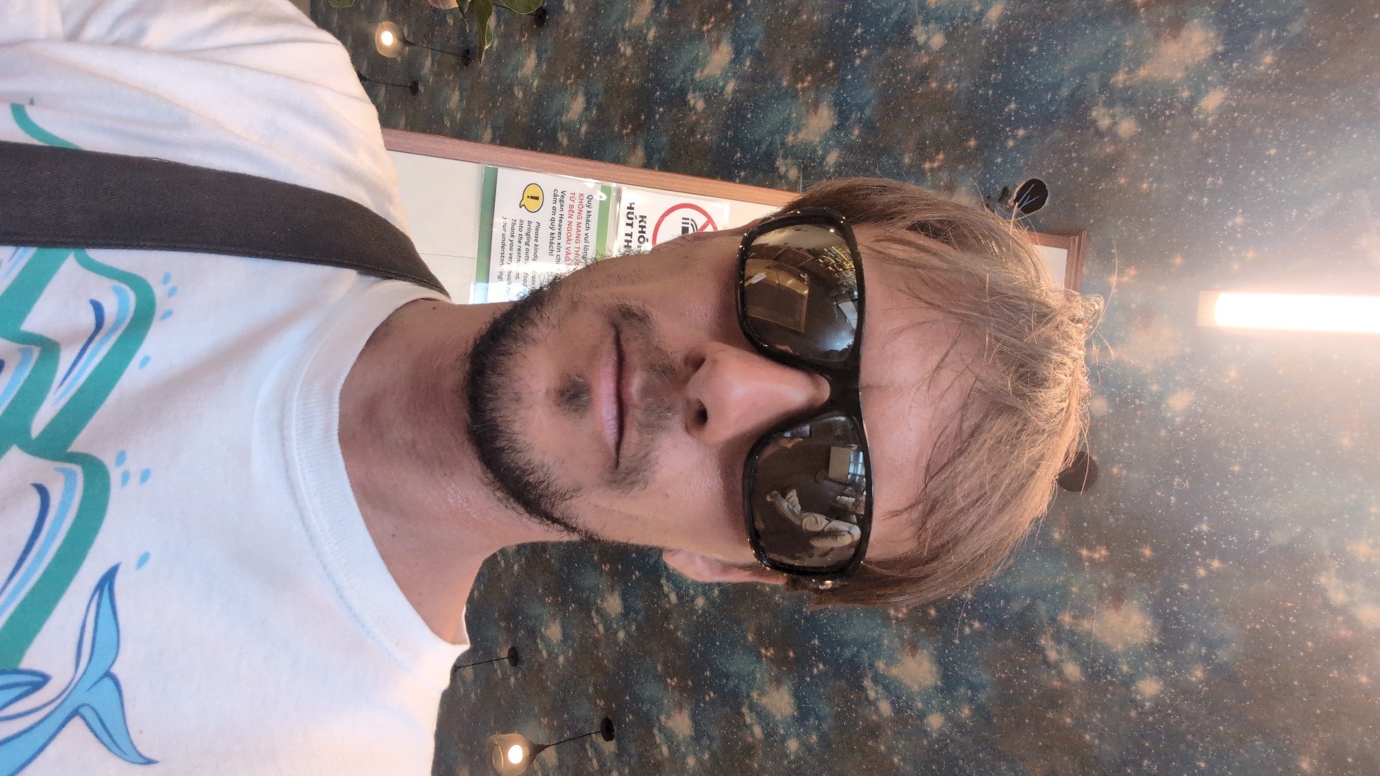 A man wearing sunglasses and a white t-shirt with a whale design is taking a selfie indoors. He has short brown hair and a goatee. He is standing in front of a wall with a galaxy-themed wallpaper and a sign that reads "Please kindly refrain from bringing out food into the room". The sign appears to be bilingual, with Vietnamese text above English. The man is carrying a dark strap over his left shoulder. The lighting is dim, with a bright strip light above and two pendant lights visible in the background. The scene is likely in Da Lat, Vietnam, given the Vietnamese text on the sign.