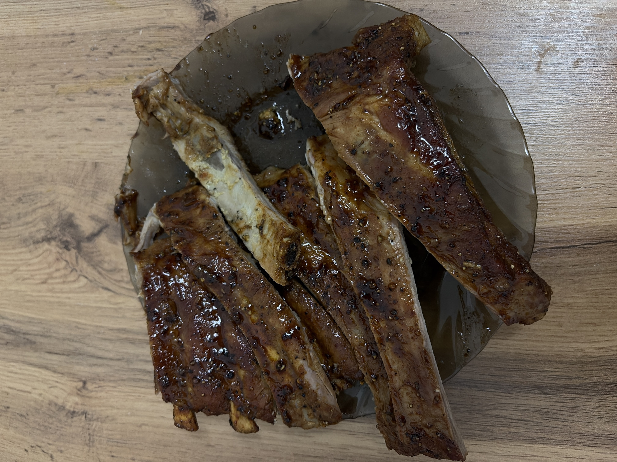 The image shows a plate of ribs on a wooden table. The ribs are glazed with a dark sauce, possibly barbeque or teriyaki. Some of the ribs have been eaten, revealing the bone. The plate is a translucent dark color. There are no people visible in the image. The location in Chaikovsky, Russia is not evident from the image.