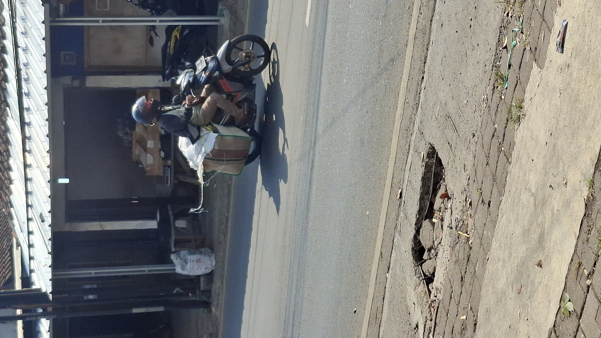 A man rides a motorcycle loaded with cargo down a street in Cilaku, Indonesia. The man, wearing a helmet and casual clothing, is positioned centrally in the frame, and the motorcycle is in motion. The cargo, consisting of sacks and boxes, is secured to the rear of the motorcycle. The background features buildings and a roadside with a damaged sidewalk in the foreground. The lighting suggests a sunny day, casting shadows on the road.