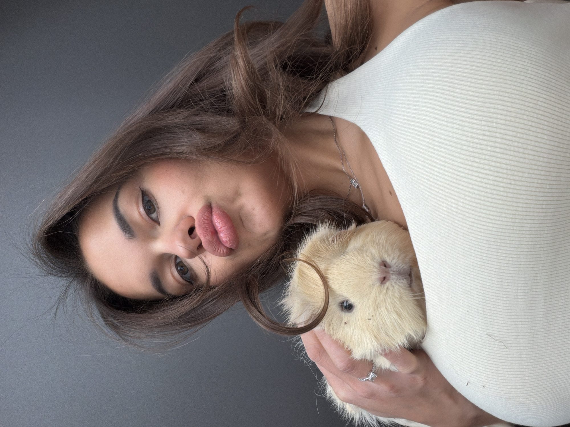 A woman is holding a guinea pig. The woman has brown hair, fair skin, and wears a white top and silver necklace. She is making a kissy face toward the camera. She wears a ring on her left ring finger. The guinea pig is cream-colored. The background is a solid gray. There is no indication of Krasnoyarsk, Russia, or any other specific location.