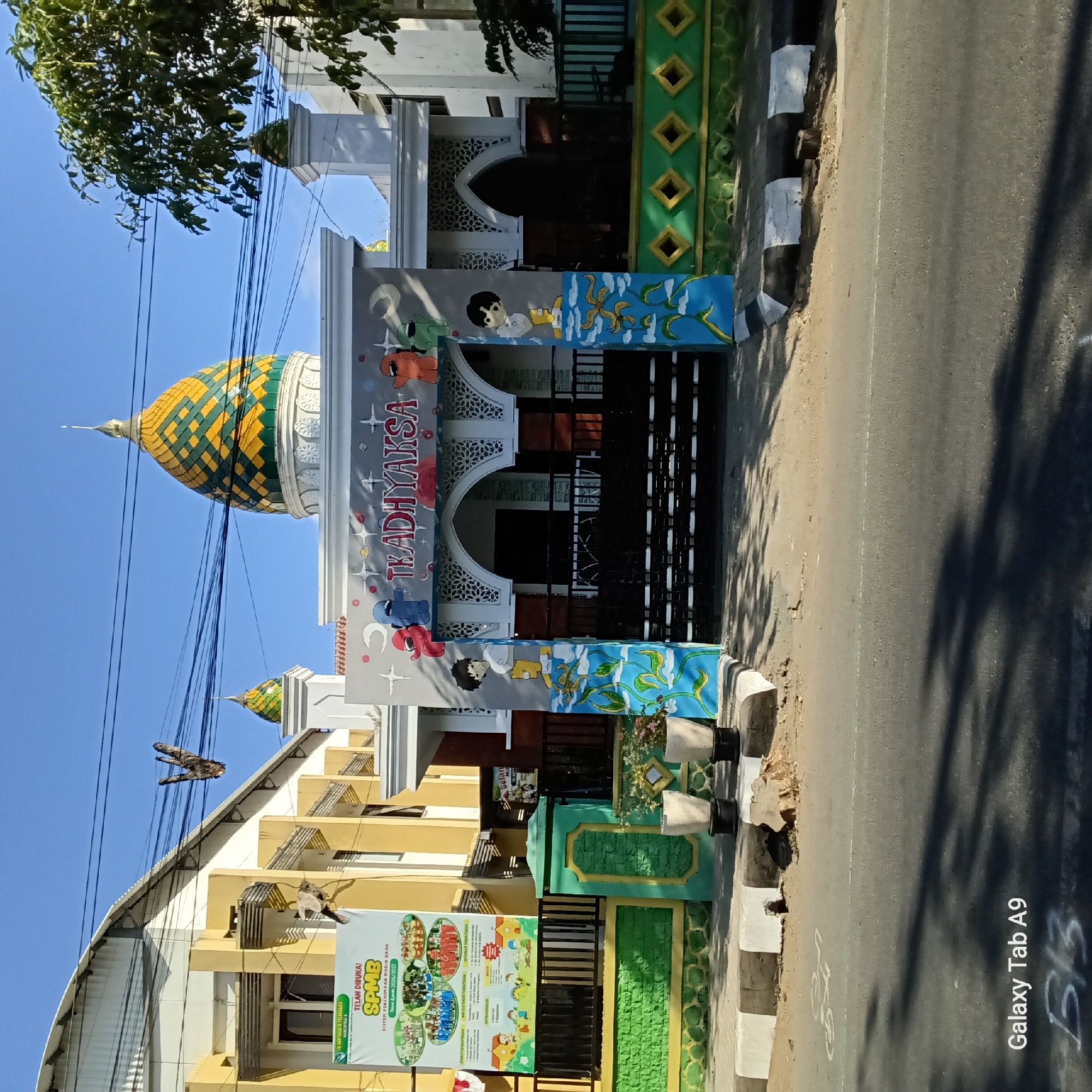 The image shows the entrance to TK Adhyaksa in Trenggalek, Indonesia. The building features a prominent, colorful dome and decorative arches. The entrance gate is adorned with murals of cartoon characters. A banner advertising the school is displayed on an adjacent building. The scene is viewed from a street, with sidewalks and a road in the foreground. Two potted plants are placed near the gate. The sky is clear and blue.